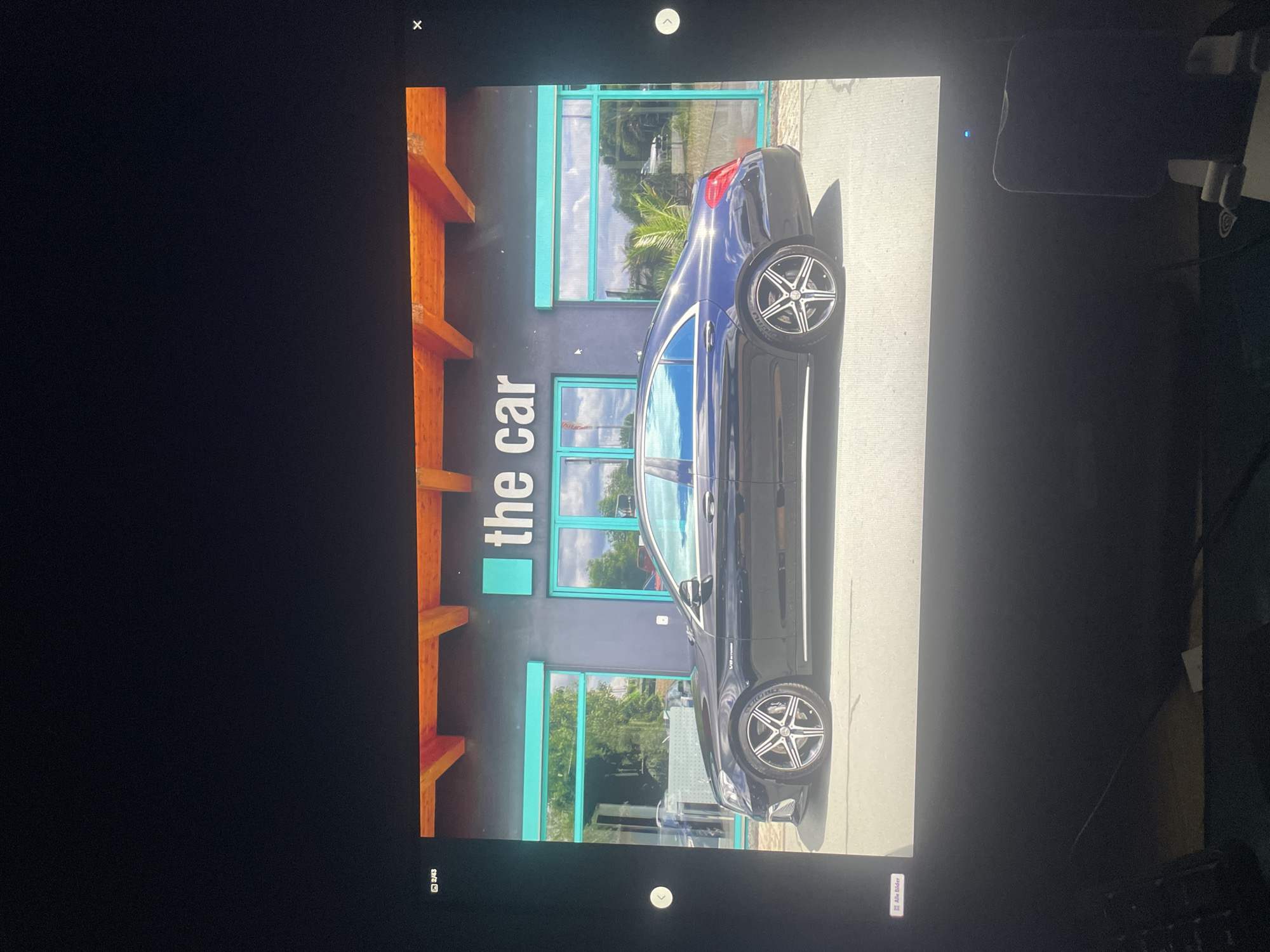 A black Mercedes-Benz sedan is parked in front of a building with the words "the car" displayed on its facade. The building has turquoise-colored window frames and wooden beams supporting an overhang. Trees and clouds are reflected in the windows. The car's rims are silver and black, and the tire sidewalls show the brand "Pirelli." The scene appears to be outdoors, likely a dealership or showroom, under a bright sky. The image is a photograph displayed on a screen, with UI elements such as navigation arrows and a thumbnail preview visible at the edges.