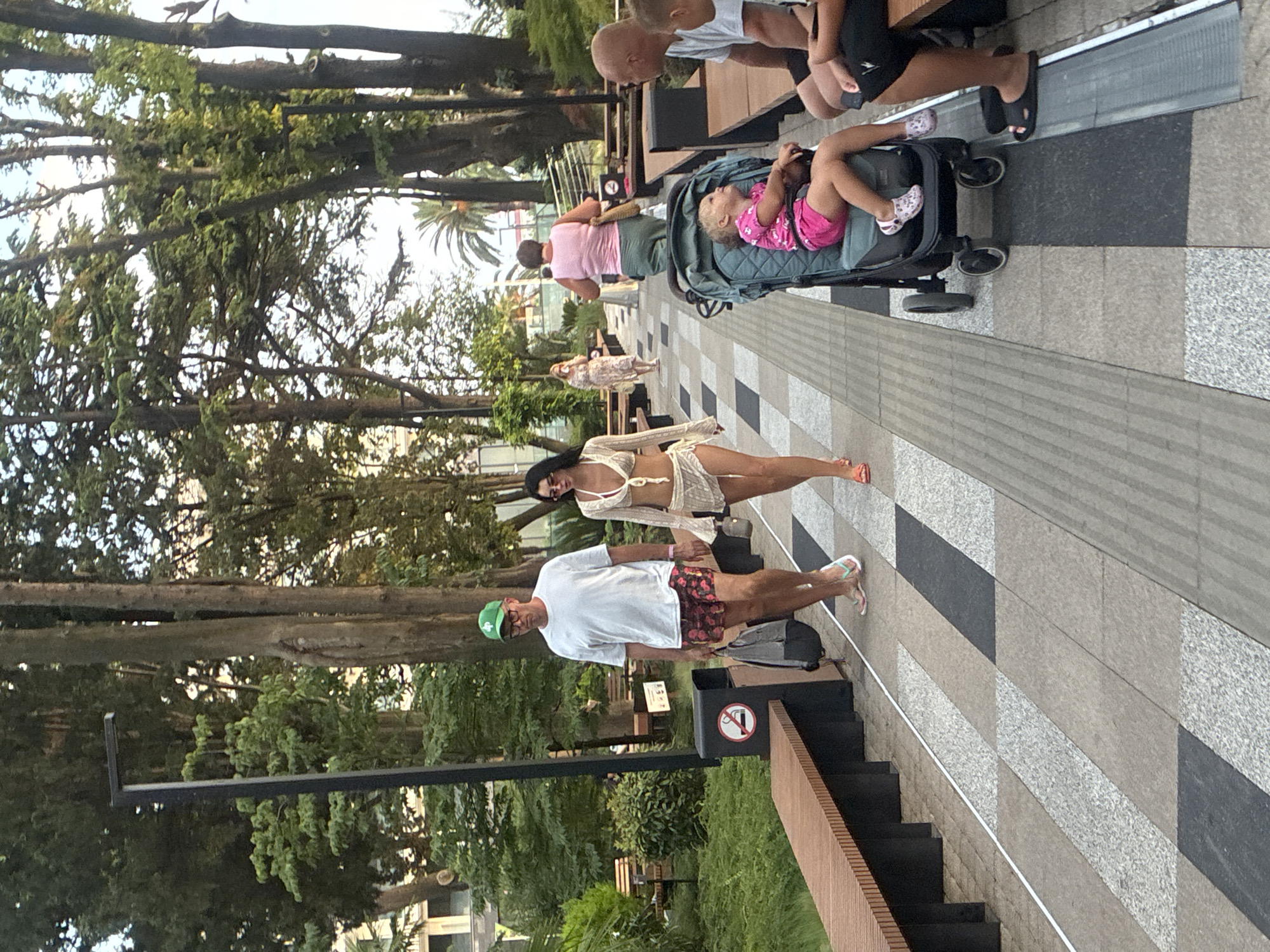 The image depicts a public walkway in Sochi, Russia, likely a park or promenade. The walkway is paved with a patterned arrangement of light and dark stone tiles, with a raised edge. Along one side of the walkway, there are modern wooden benches with dark metal supports. A trash receptacle with a no-smoking sign is visible.
Several people are present:
* A man and a woman are walking towards the viewer. The man wears a white t-shirt, floral patterned shorts, a green baseball cap, and glasses. He is carrying a dark backpack. The woman wears a light-colored, open-knit two-piece outfit and sunglasses.
* Further down the walkway, a woman in a pink tank top and green pants walks away from the viewer, carrying a straw bag.
* To the right, a man and a child are seated on a bench. The man wears a white t-shirt and black shorts.
* A toddler is seated in a teal stroller positioned near the seated man. The toddler wears a pink outfit.
* In the background, other individuals and palm trees are visible, suggesting a warm climate. The overall environment is lush with trees and appears to be a pleasant outdoor space.