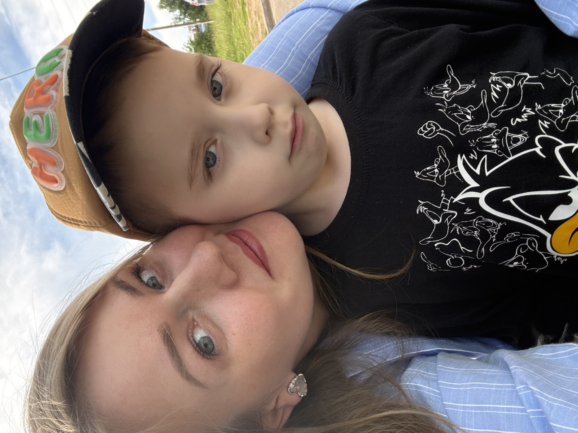 A close-up, over-the-shoulder selfie features a woman with blonde hair and a blue and white striped shirt, her face positioned close to a young boy wearing a brown baseball cap with "HERO" spelled out in colorful, raised letters and a black t-shirt with a cartoon duck print. The woman has blue eyes and is wearing a heart-shaped earring. The boy has blue eyes and a slightly downcast expression. They appear to be outdoors, with a cloudy sky and greenery in the background. The location is identified as Chaikovsky, Russia, likely an outdoor recreational area or street.