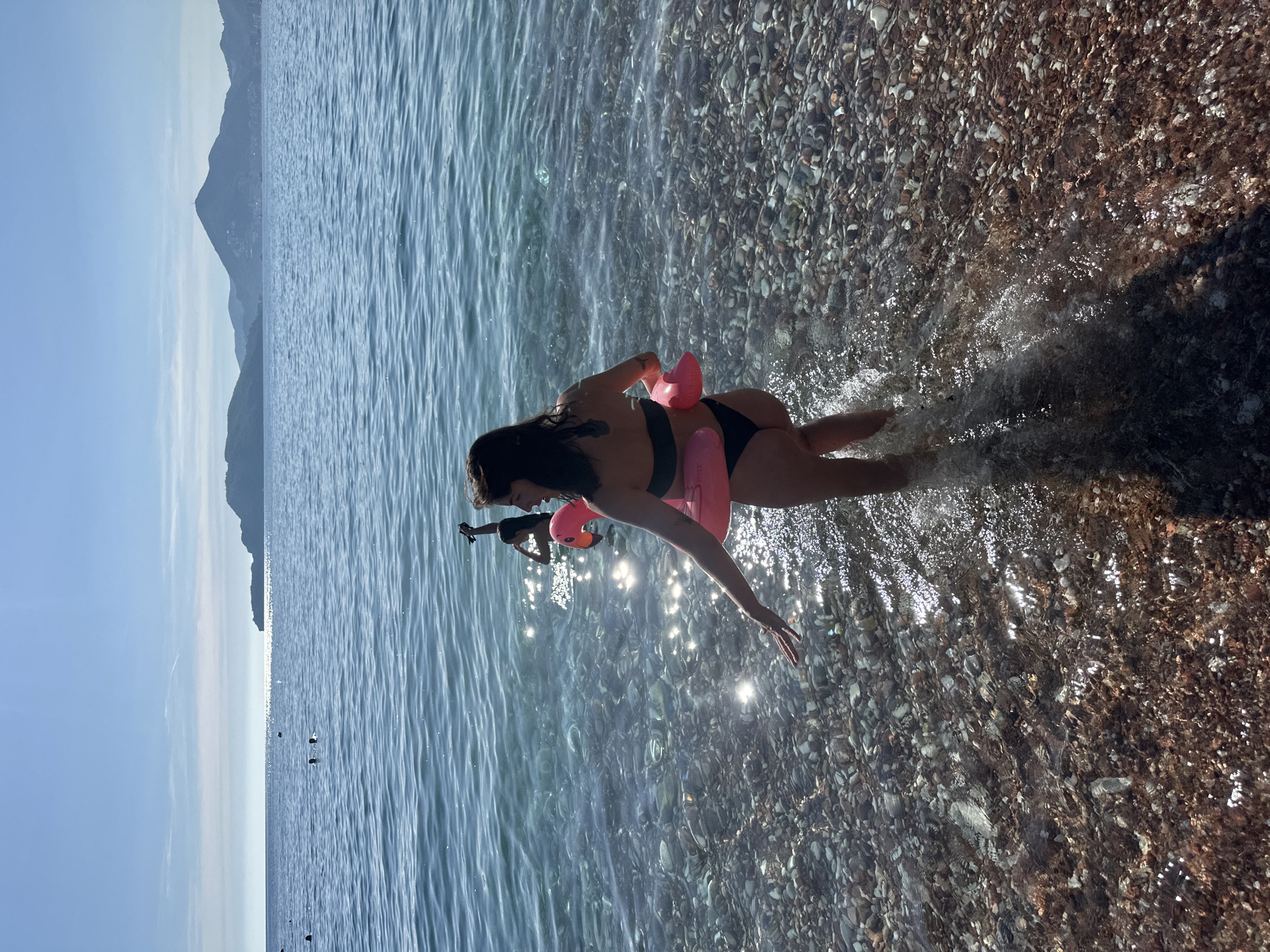 The image shows a person in the water at a pebbly beach. The person is wearing a black bikini and a pink flamingo-shaped inflatable. The sun is shining brightly, creating reflections on the water. In the background, mountains are visible, suggesting a coastal location. The water is clear and shallow, allowing the pebbles on the seabed to be seen. The image is oriented vertically. The scene appears to be a leisure activity on a sunny day.