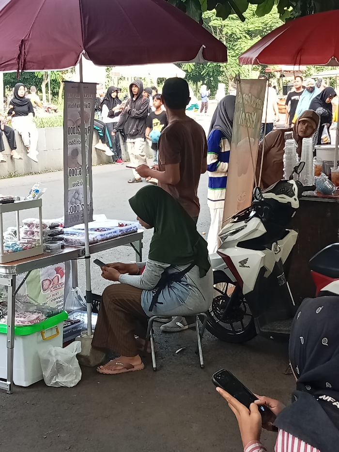 This image captures a vibrant outdoor market scene in Singaparna, Indonesia, likely during the daytime under bright, partially shaded conditions. Numerous people are gathered around various stalls, suggesting a social or commercial hub.
In the foreground, a woman wearing a dark green hijab and brown pants is seated on a small metal stool, engrossed in her mobile phone. Her light-colored sneakers are resting on the stool beneath her. To her left is a vendor stall featuring shelves displaying small, round items in clear containers, possibly sweets or snacks. A tall, narrow sign on this stall has partially visible text including "Chill Bites", "Dalgona", and pricing like "000.S" and "00.000". Beneath this, a green and white cooler and plastic bags are visible.
To the right, a white Honda scooter is parked near another stall. Behind this stall, a woman in a brown hijab peers out, looking towards the camera, while plastic cups and bottles, indicative of cold drinks, are arranged on the counter. A man in a brown t-shirt is standing near this stall, his back to the camera. Further back, on a low concrete wall, a woman in a black hijab and white attire is seated, observing the surroundings. Other individuals, many also wearing head coverings, are seen milling about in the background, some near other vendors, others simply standing or walking. Lush green trees provide a natural canopy overhead. Another person in the far foreground has a phone in hand, and "NFA" is faintly visible on their clothing. The overall atmosphere appears relaxed and bustling with daily activity.