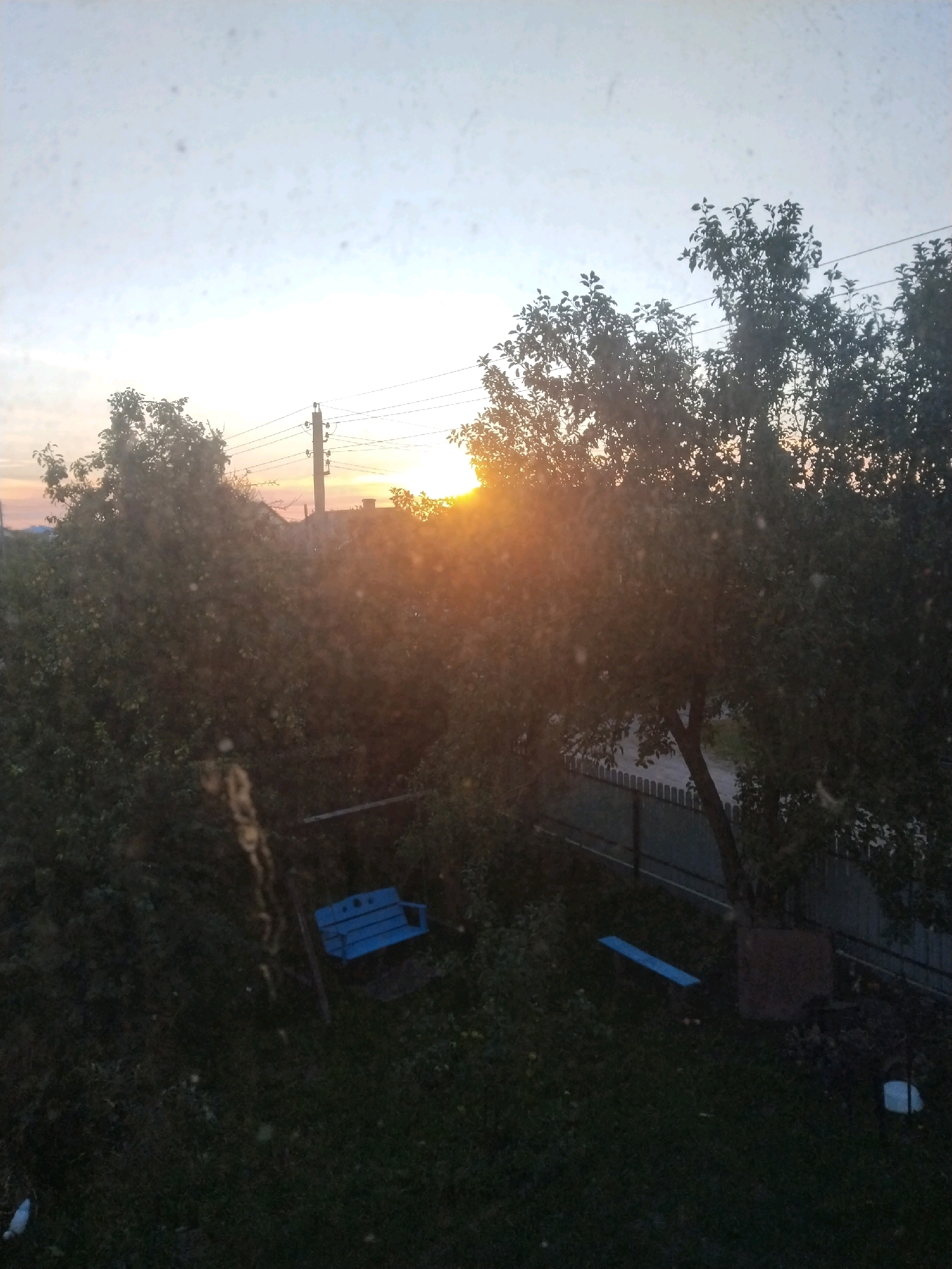 This image depicts a backyard scene at dusk in Pyetrykaw, Belarus, viewed through a window with some dust or smudges on the glass. The setting sun casts a warm glow through the trees, silhouetting their branches and leaves. In the foreground, a dark green lawn is visible, with a blue swing set and a light blue plank lying on the grass. A metal fence borders the property in the background. Power lines and a utility pole are visible against the pale sky, suggesting a residential area within the city. The overall impression is one of a quiet, rural-like evening in a Belarusian town.