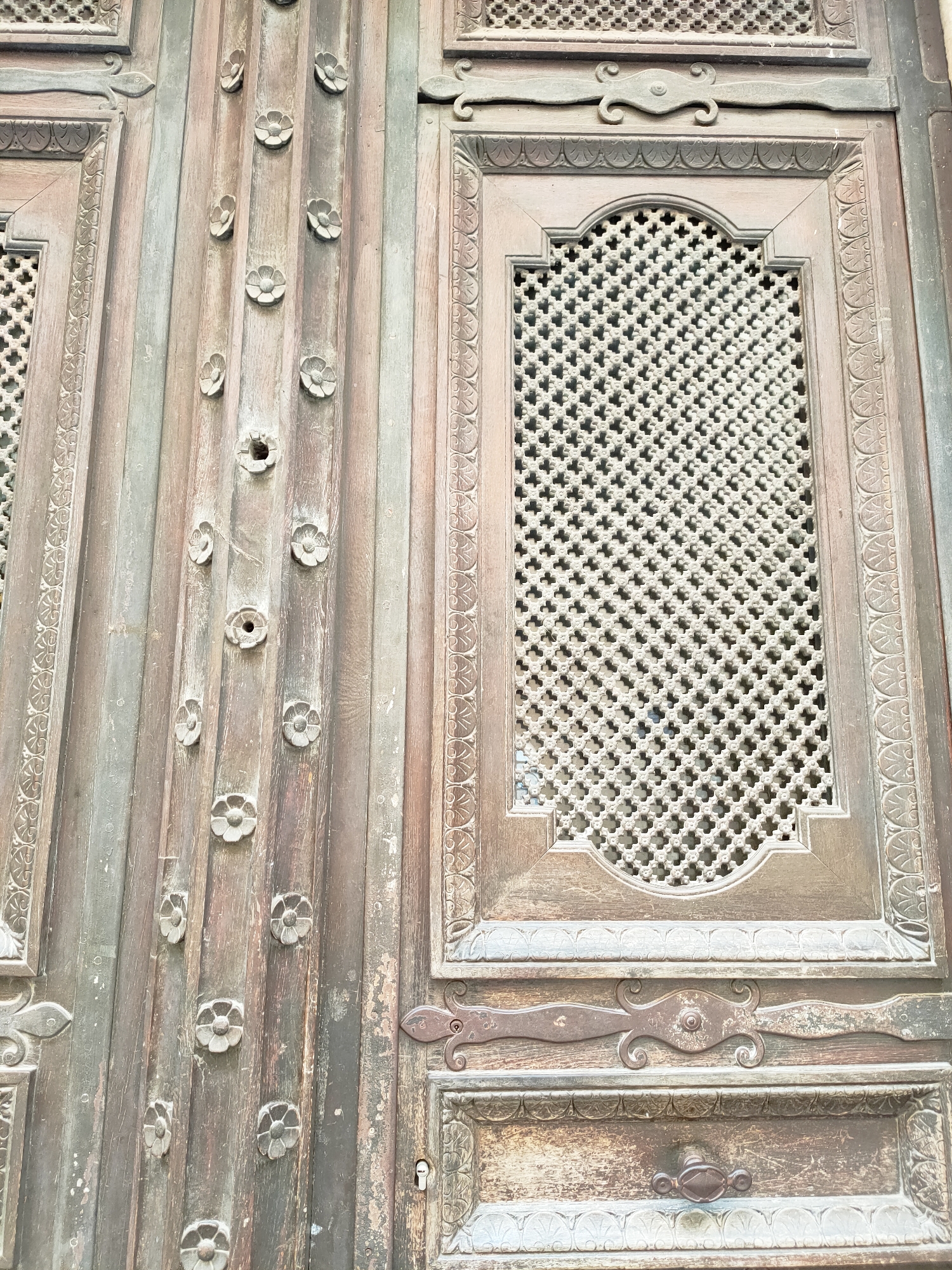 The image shows a close-up of an old, weathered wooden door with intricate carvings and metal hardware. The door appears to be composed of vertical wooden planks. Small, circular floral carvings are spaced out along some of these planks. One section of the door features a decorative metal lattice inset into a framed panel, surrounded by carved borders. Below this, there is a horizontal decorative metal element and another framed panel containing a keyhole and a drawer pull. The wood shows signs of age, with wear, discoloration, and some surface damage. The overall impression is one of historical craftsmanship and the passage of time. The location is identified as the Maison Julia, Perpignan, France.