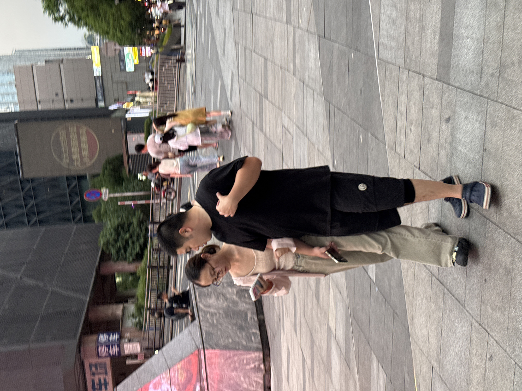 The image captures a street scene in the Yuzhong District of China. In the foreground, a couple walks together, with the woman looking at her smartphone while holding a second phone in her other hand. She is wearing a light pink cardigan over a beige top and tan wide-leg pants. The man beside her is wearing a black t-shirt and black shorts. They are walking on a paved pedestrian area with large square tiles. In the background, other pedestrians are visible, along with buildings, trees, and traffic signals. One building has a large screen displaying an advertisement. The overall environment appears to be a busy urban area.