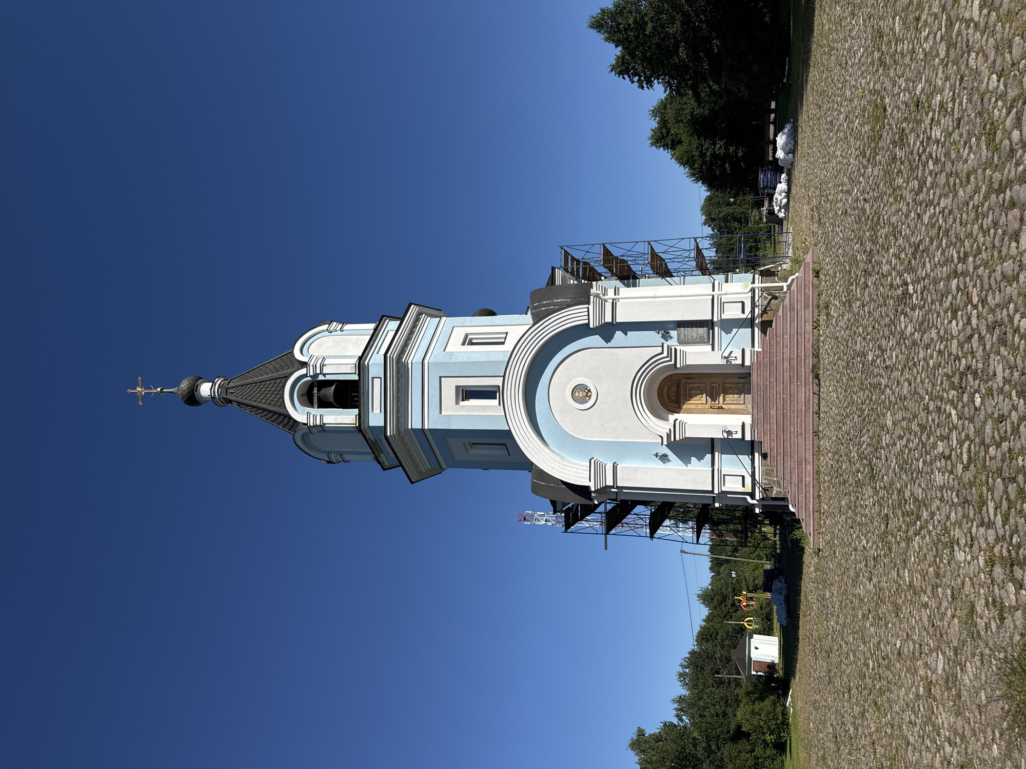 The image shows the Church of the Kazan Icon of the Mother of God in St. Ozerki, Russia, on a clear day. The church is a multi-tiered structure with a light blue and white facade, topped by a dark dome and a golden cross. Scaffolding is erected on the left side of the building, suggesting ongoing construction or renovation. The church is set on a cobblestone plaza, with a set of red-carpeted stairs leading to the entrance. To the left of the church, a small white building with a red roof is visible. The background consists of green trees and a clear blue sky. There are no people or animals visible in the image. Several white bags, possibly filled with materials, are piled on the ground to the right of the church.