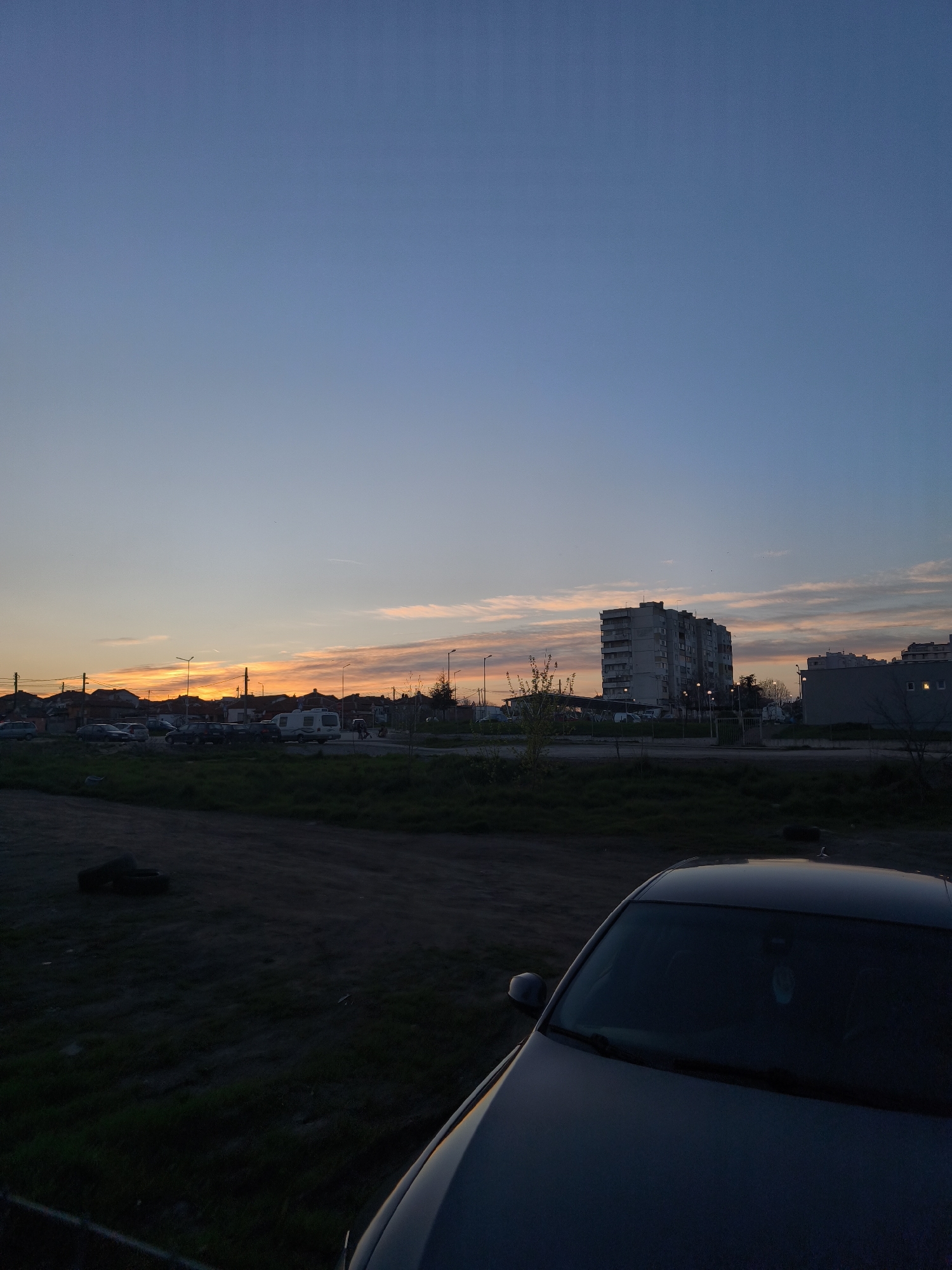 The image depicts an urban scene at dusk. The sky transitions from blue at the top to orange at the horizon, indicating a sunset or sunrise. In the middle ground, there are several buildings, including a large, grey, multi-story apartment building. Other smaller structures, possibly houses, and streetlights are also visible. A line of trees and bushes runs along the roadside. A parked car occupies the foreground. The overall environment suggests a residential area in a city or town. Based on the urban landscape featuring Soviet-era apartment blocks and the general architectural style, the image could be taken near Burgas, Bulgaria.