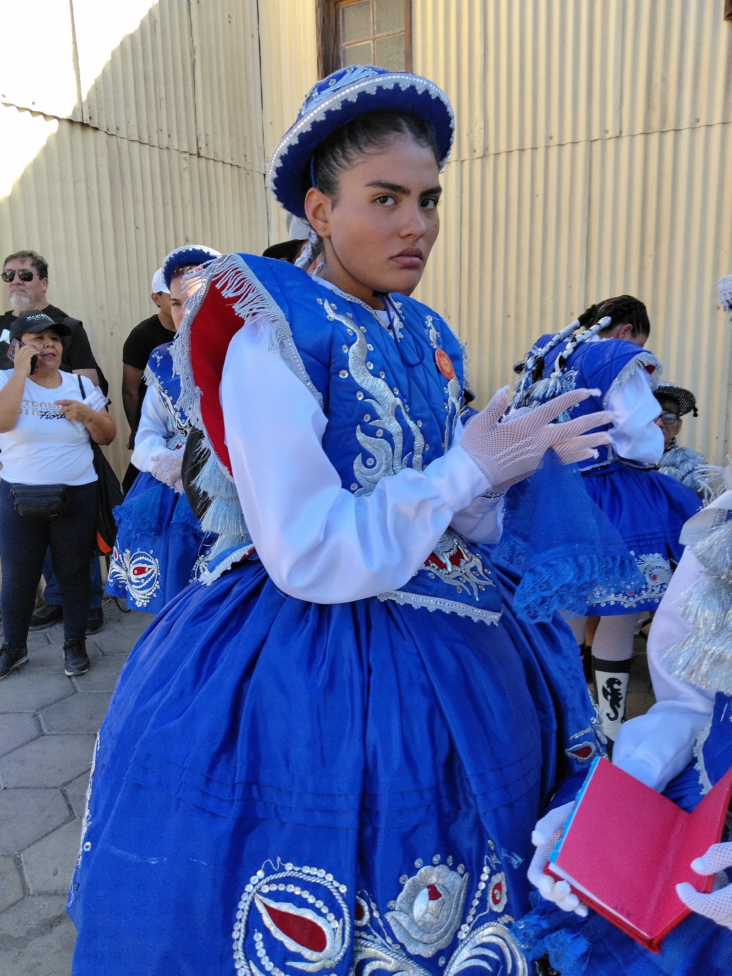 In Pozo Almonte, Chile, a young woman in a vibrant blue and white traditional costume with silver embroidery is the central subject of this image. She wears a matching blue hat adorned with white trim and silver detailing. Her costume includes a voluminous blue skirt and a fitted blue bodice with white sleeves. She is also wearing white lace gloves. Her expression is serious as she looks directly at the camera, holding a small, open red book in her gloved hands.
Behind her, other individuals are visible, also dressed in similar colorful traditional attire, suggesting a cultural celebration or parade is underway. Some wear matching blue hats and ornate costumes with red and white accents. One person in the background has their hair in prominent braids. The setting appears to be outdoors, with a corrugated metal wall forming the backdrop. The ground is paved with cobblestones. The lighting suggests it is daytime, possibly late afternoon due to the long shadows cast on the wall. The overall atmosphere seems festive, given the elaborate costumes and the gathering of people.