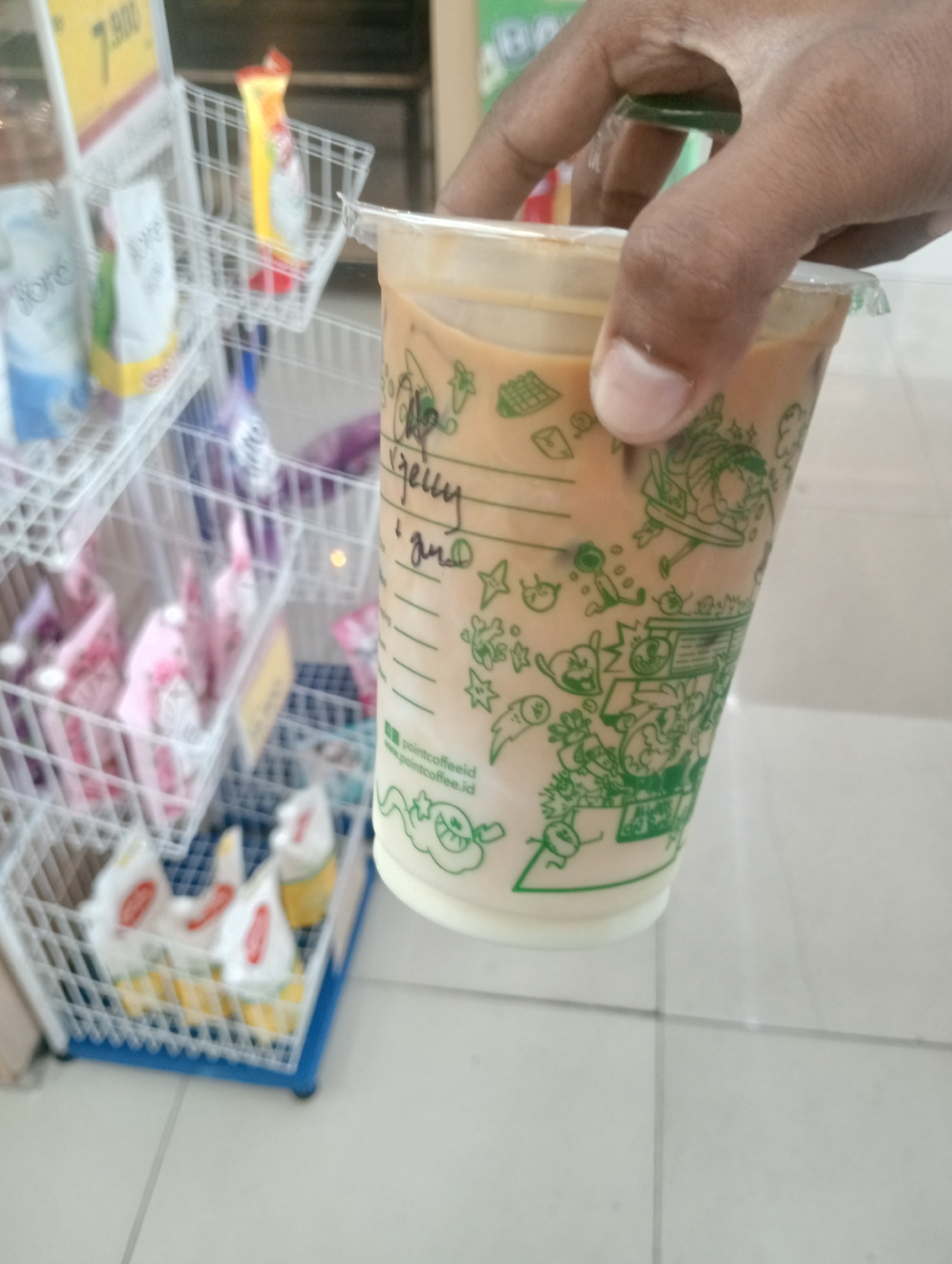 An overhead shot shows a person's hand holding a plastic cup with a light brown beverage and white cream on top. The cup has green cartoon-like drawings on its exterior. Behind the cup, several white wire baskets are filled with various products. The basket to the left holds packaged goods with blue and white labels, while the middle basket contains items in yellow and red packaging. The basket on the right also holds yellow and red packaged items. In the background, a yellow sign with dark lettering is visible. The floor appears to be tiled. The scene is likely within a retail store or convenience store in Surabaya, Indonesia.