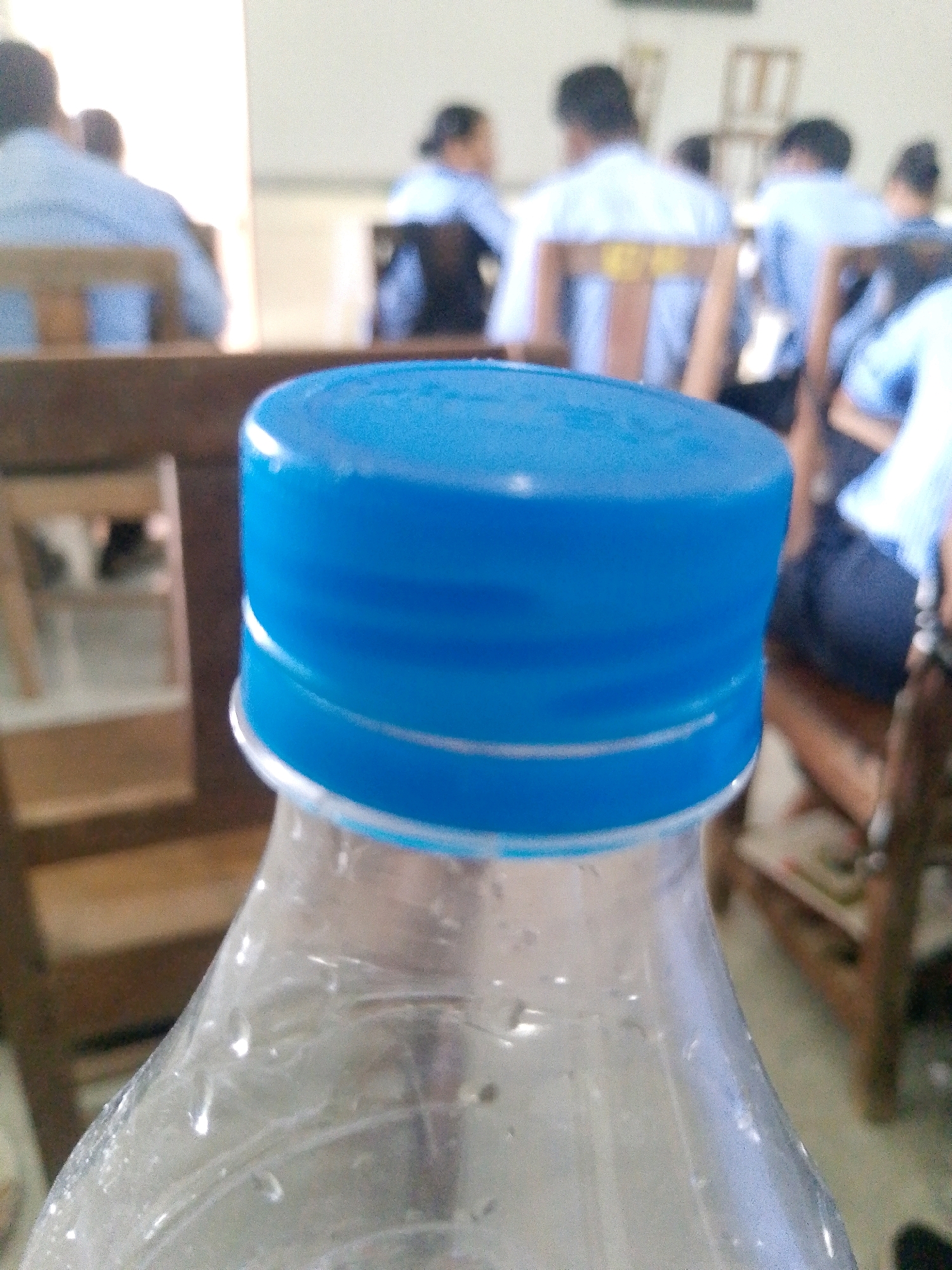 The image shows a close-up of a blue plastic bottle cap in the foreground. The cap is slightly out of focus, with the bottle itself being more in focus. The background is blurred and depicts an indoor scene with several people seated at tables. The people appear to be students or attendees in a classroom or lecture hall setting. Some individuals are wearing light-colored shirts, and others are in darker attire. Wooden chairs and tables are visible. The environment suggests a public or educational institution. No specific details about Sitarampuram, India, are discernible from the image itself.