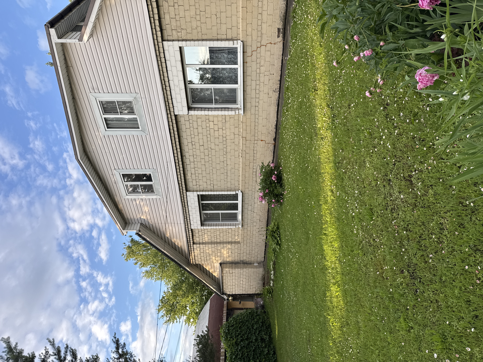 The image shows a side view of a two-story house with light beige brick on the lower level and light gray vinyl siding on the upper level. The house has several windows; the lower level has one large window with white trim, and the upper level has two smaller windows with white trim. A green lawn with scattered white and pink flowers covers the foreground. A bush of dense green foliage is visible on the left side of the frame, partially obscuring a section of fencing and a sloped roof. In the background, trees and a blue sky with scattered white clouds are visible. The overall scene depicts a residential property on a sunny day. The specific location is a residential area in Naro-Fominsk, Russia. No people or actions are depicted in the image.