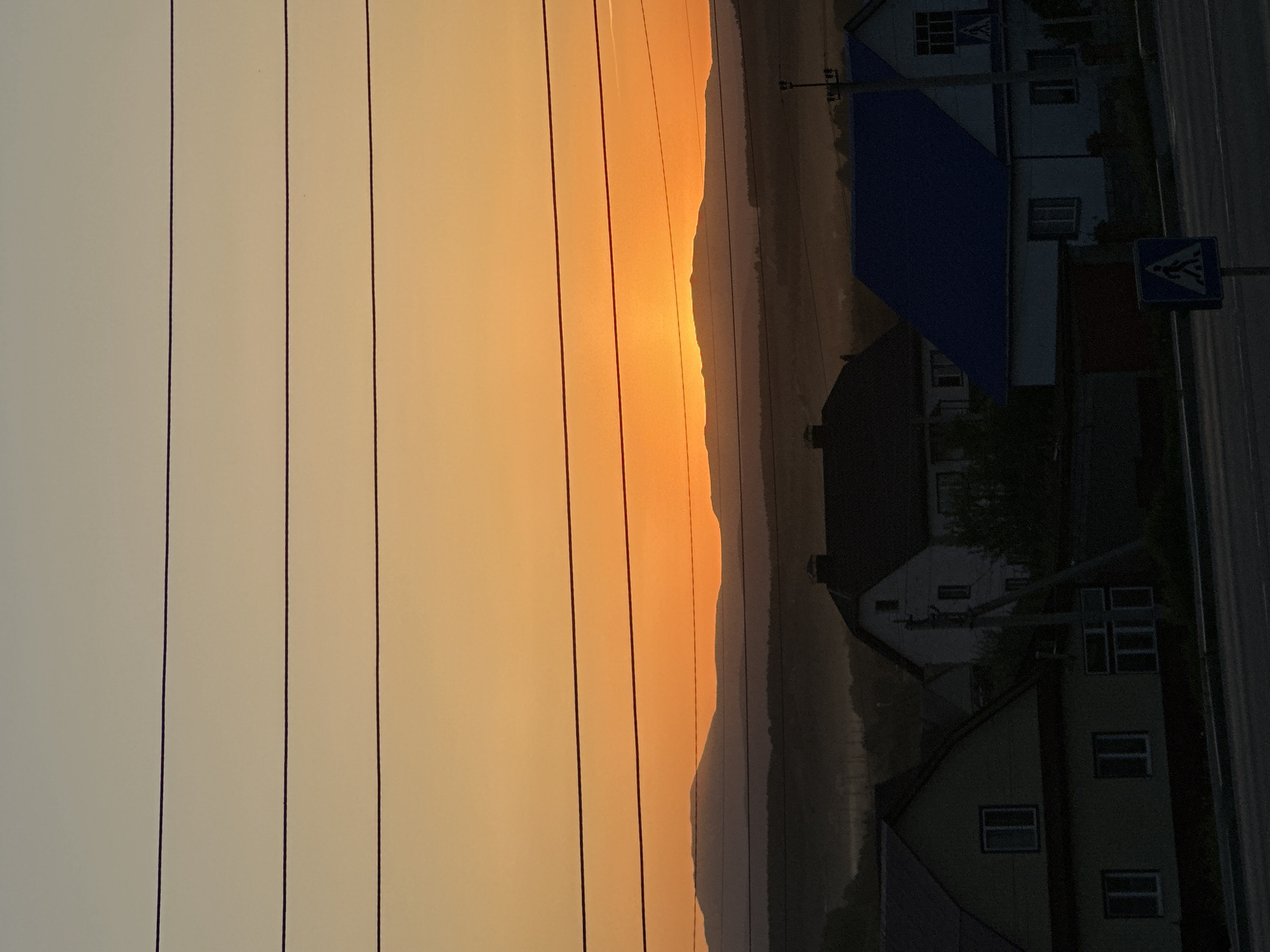 A landscape photograph depicts a scene with an orange sky on the left, likely a sunset or sunrise. Several power lines run vertically down the left side of the image. To the right of the sky and power lines, a mountainous region or hills are silhouetted against the bright light. In the lower right, there are several buildings and a road with a pedestrian crossing sign. The image is taken in a city where the location is unknown.