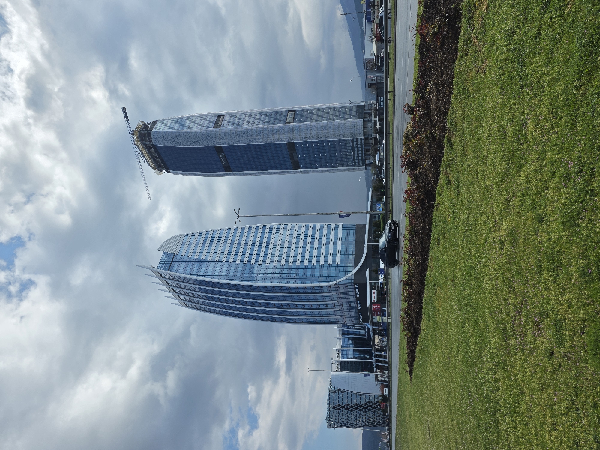 The image depicts two modern skyscrapers in Sofia, Bulgaria, set against a cloudy sky. One building is still under construction, with a crane visible at its apex. The other completed skyscraper showcases a curved, glass façade. In the foreground, there is a strip of green grass and a road with sparse traffic. Building signs for "Pasadow" "DISCORDIA" and "KARE" are visible. The architectural styles of the buildings are modern and futuristic.