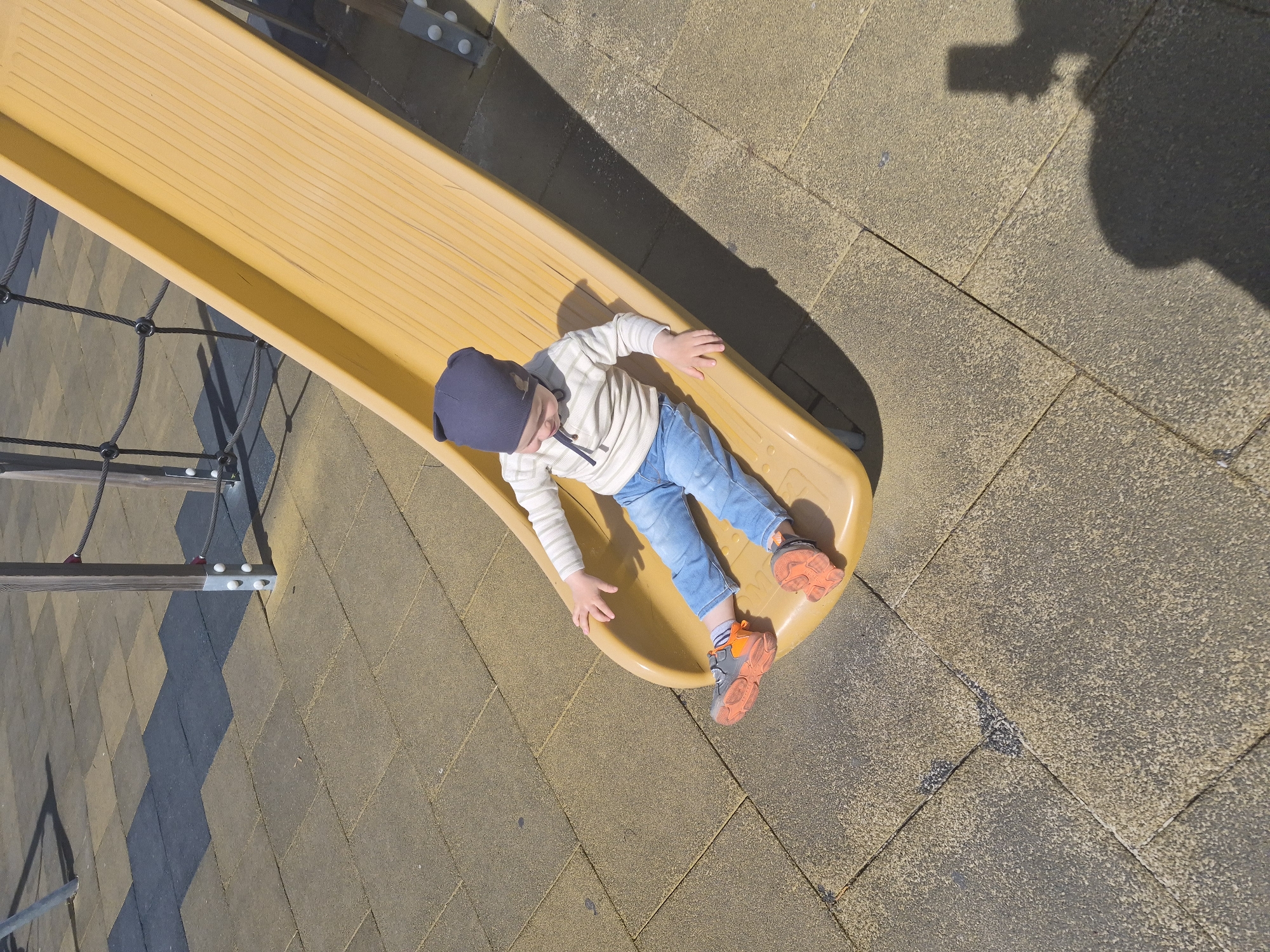A young child, wearing a dark blue beanie, a striped long-sleeved shirt, light blue jeans, and orange and grey sneakers, is lying on their back in a yellow slide. The slide is located on a playground with a tan paved surface. A shadow is cast on the ground, possibly of the photographer. The location is not identifiable as Tobolsk, Russia from the image.