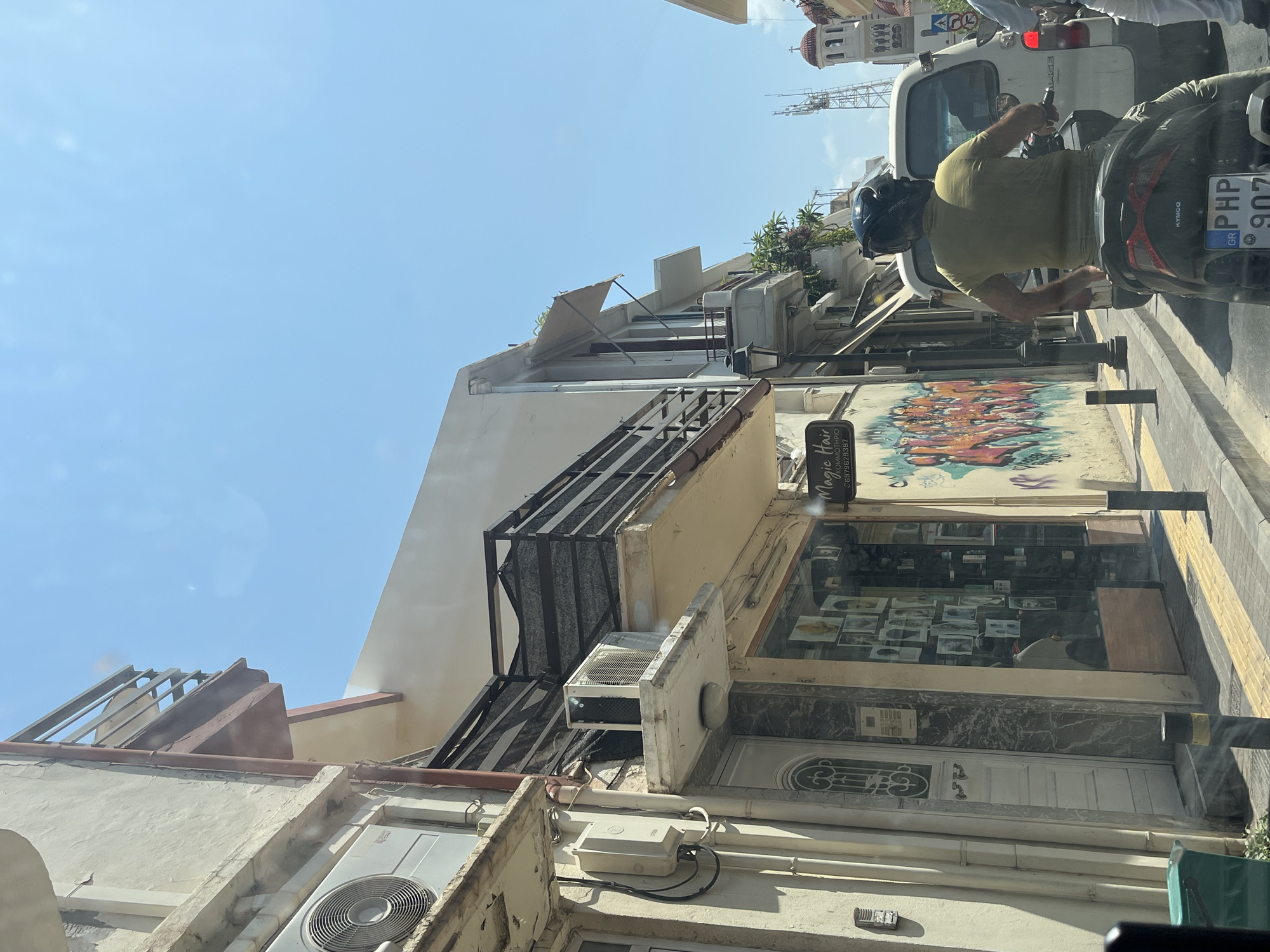 The image captures a street scene in Rethymno, Greece, under a clear blue sky. The perspective is from a vehicle, looking towards the right side of the street. On the left, a building features an outdoor air conditioning unit mounted on a balcony. Below, a shop window displays photographs and products. A white door with decorative elements is next to the shop. Graffiti art adorns the wall to the right of the shop.
In the foreground and middle ground, a person wearing a yellow t-shirt and a black helmet is riding a scooter, moving away from the viewer. The scooter has a license plate visible with Greek markings. Behind the scooter, a white van is partially visible. Further down the street, a tall telecommunications tower and the domed roof of a building, likely a church, can be seen. The street is paved with sidewalks marked by yellow and gray stripes.