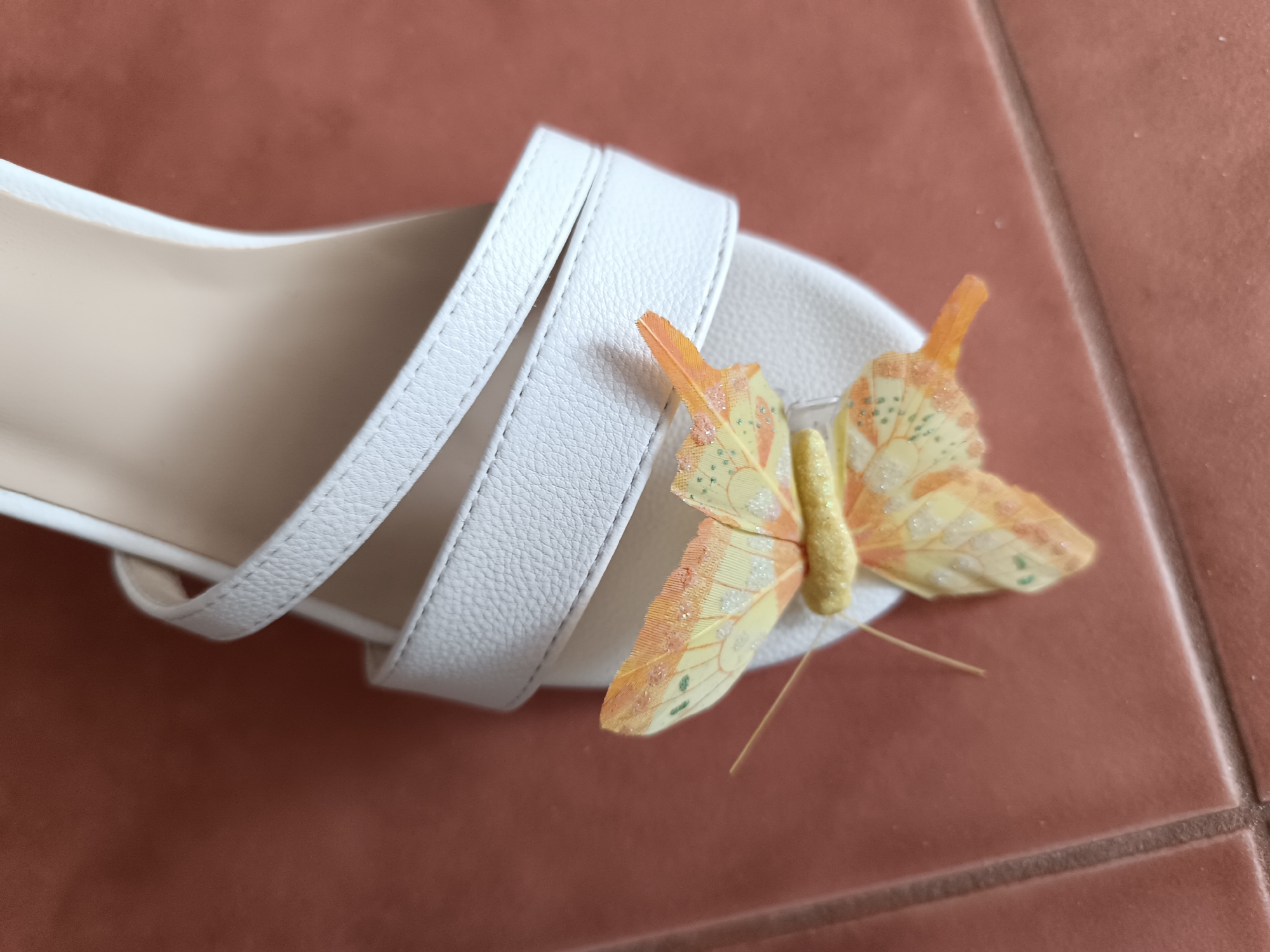The image shows a close-up, overhead view of a white sandal with decorative straps and a small, artificial butterfly embellishment. The sandal is placed on a tiled surface with a reddish-brown hue. The butterfly is yellow and orange with glittery accents and appears to be made of fabric or paper. The scene is brightly lit, suggesting it was taken during the daytime, likely indoors with natural light. There are no people visible, and the focus is solely on the sandal and its decoration. The background of the tiles is plain, with no discernible text or further location cues beyond the tiled surface. The overall impression is that of a product shot or a detail of an accessory.
