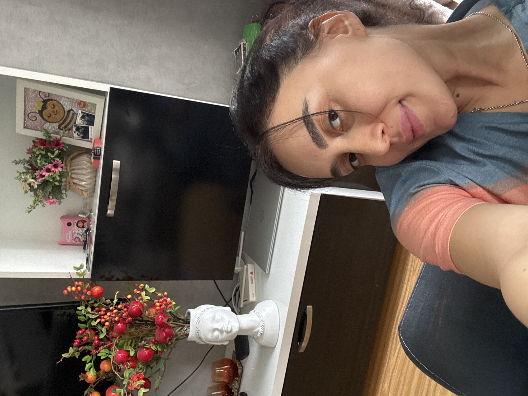 A woman is captured in a close-up, angled shot, appearing to take a selfie indoors. She has dark hair tied back, dark eyebrows, and a faint mole on her neck. Her head is tilted, and she looks directly at the camera with a subtle, closed-mouth smile. She wears a blue t-shirt with a peachy-orange tie-dye pattern on the sleeve and a thin silver chain necklace.
The background reveals a domestic setting, likely a living room or study. To her left, a white shelving unit holds a black television screen. On a shelf above the TV, a framed picture depicts a cartoon bee in an embroidered style, with two small black and white photos tucked into its corner. Below this, a vase with artificial pink and white flowers is visible, and further down, a small pink object sits on another shelf.
To her right and below her, a white table surface is seen, holding an open silver laptop and a white router/modem with connecting wires. A distinctive white decorative vase, shaped like a classical bust, is filled with artificial red berries and small pomegranates or apples. Next to it are two amber-colored glass items. The lower part of the image shows a dark brown cabinet with a silver handle and a light brown wooden floor. The overall impression is a casual, personal snapshot within a decorated home environment.