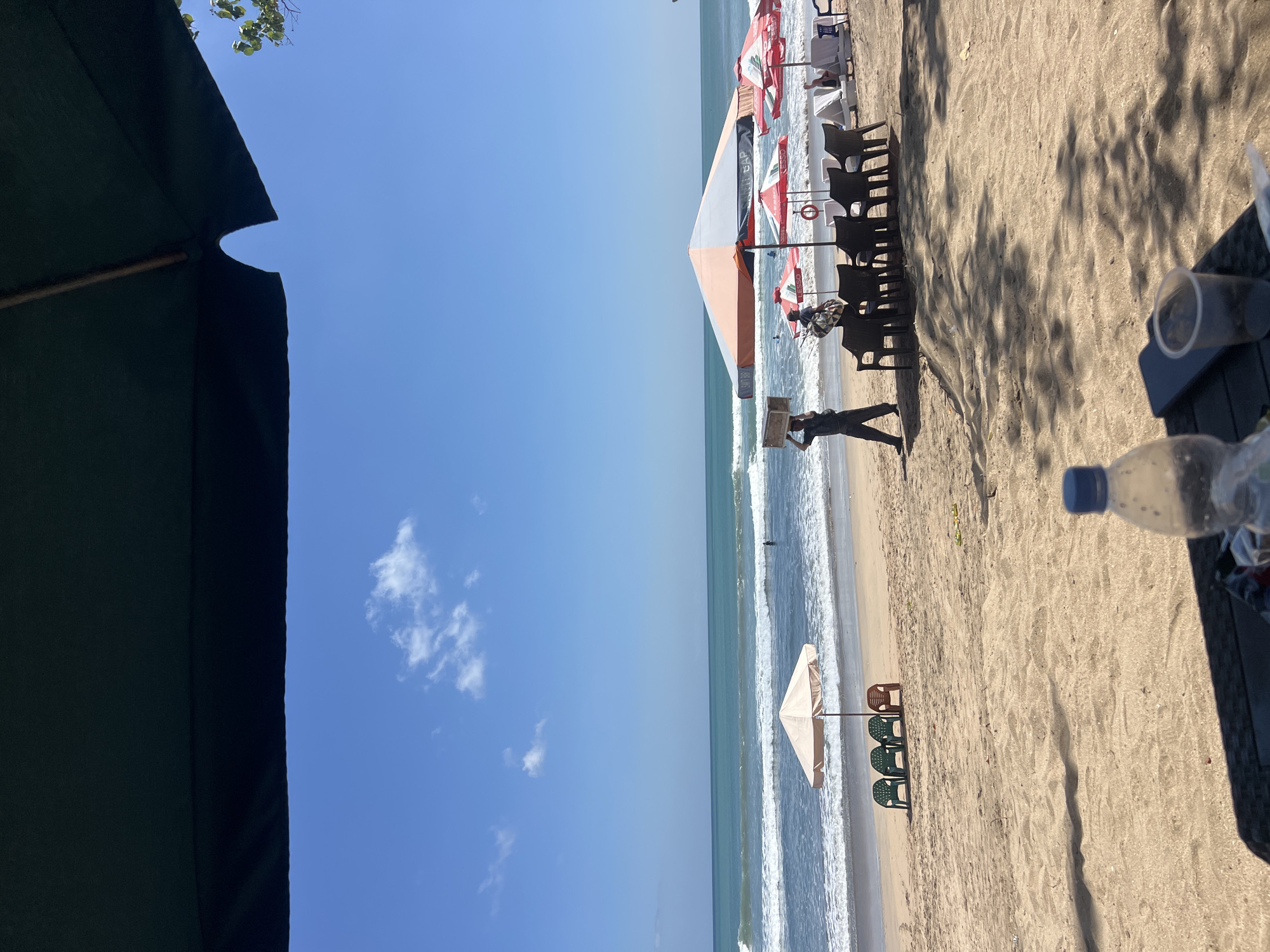 The image captures a sunny day at Kuta Beach, Indonesia, from the perspective of someone sitting under a large, dark umbrella. In the foreground, a dark surface with a plastic water bottle and a clear cup suggests a table setup. The sandy beach stretches out towards the ocean, where gentle waves roll in.
Several beach umbrellas and chairs are visible on the sand. A man in dark clothing walks across the beach carrying a rectangular wooden box on his head, while another person with a bag walks nearby. In the background, a few individuals are in the water. The sky is a clear blue with a few scattered white clouds. The overall scene depicts a typical beach day in Kuta, with a mix of leisure and activity.