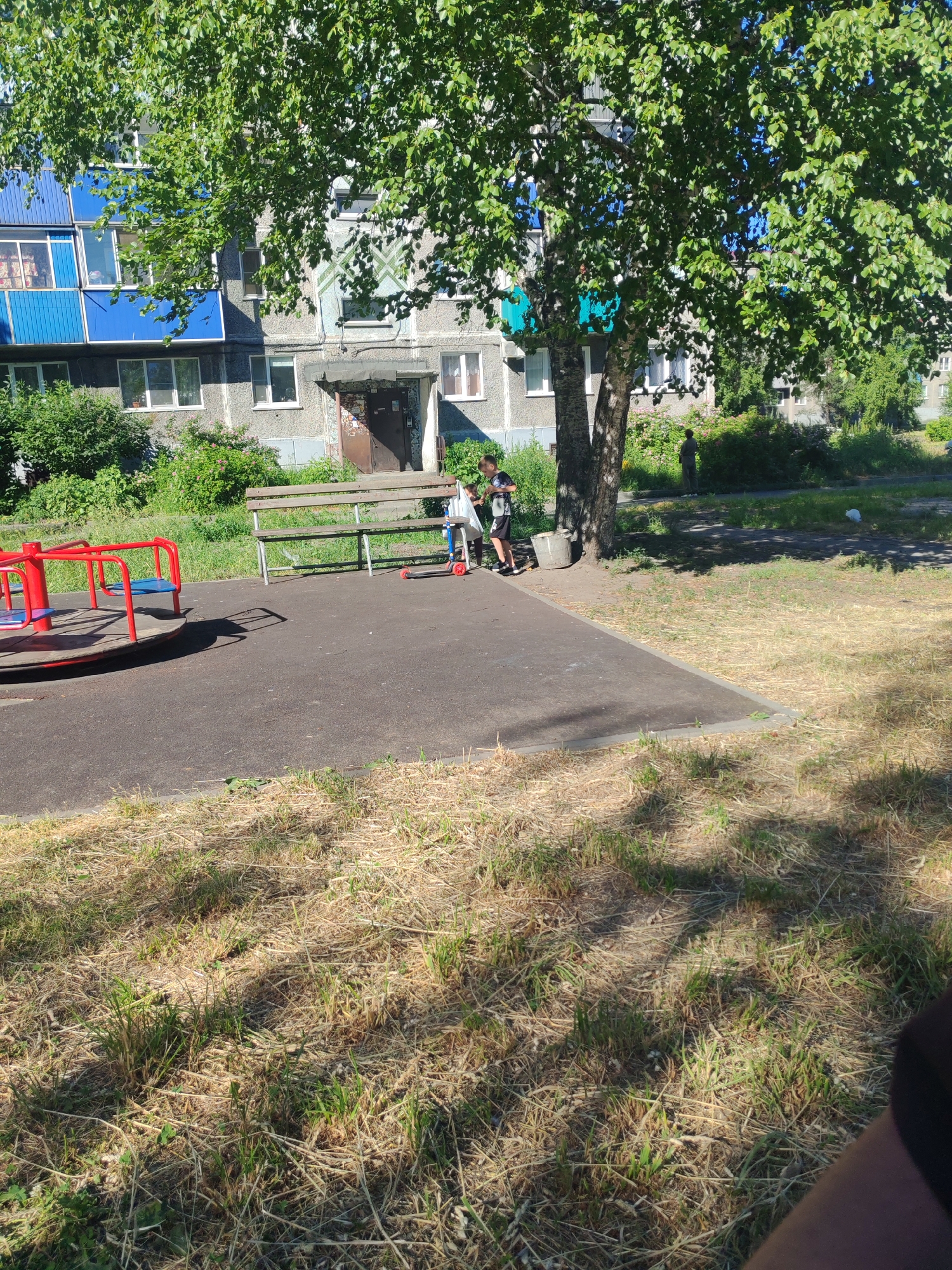 The image depicts a public playground in the city of Mezhdurechensk, Russia. A grey, multi-story apartment building, featuring blue balconies on the left side, forms the backdrop. In the foreground, a red and blue merry-go-round stands on a paved surface. To the right of the merry-go-round is a wooden park bench. Two young boys are present. One boy, wearing a white t-shirt and black shorts, stands near the bench. The other boy, in a black and white patterned t-shirt and black shorts, is on a red scooter. A bucket is visible next to the bench. A large tree with green leaves partially shades the scene. The ground is covered with dry grass and sparse green shoots.