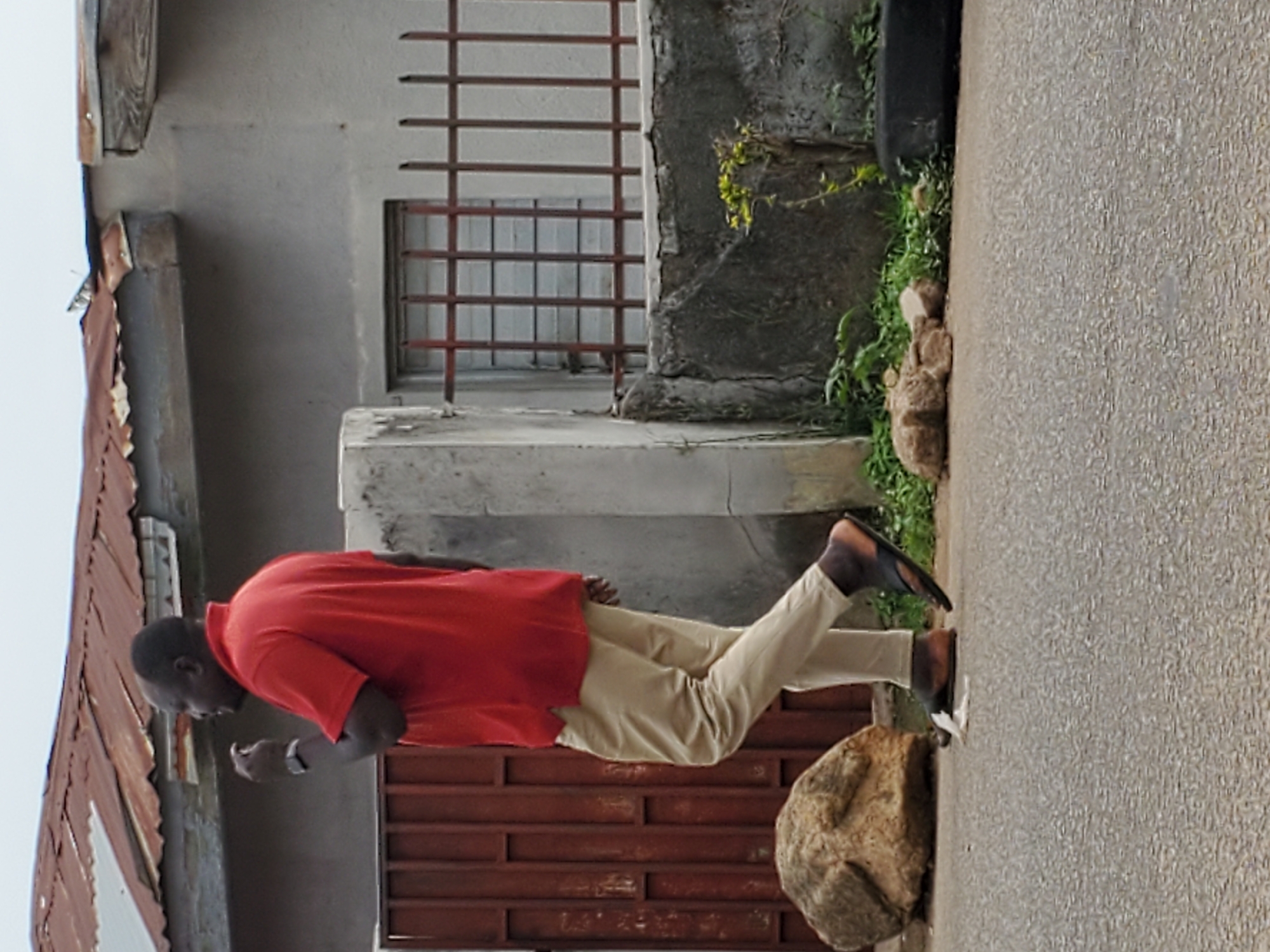 The image shows a man walking next to a building in Jos, Nigeria. He is wearing a red shirt, tan pants, and flip-flops. He appears to be looking down at something. The building has a rusty corrugated iron roof, a metal window grill, and a concrete base with vegetation growing around it. A large rock sits next to the walkway. The walkway has a textured surface.