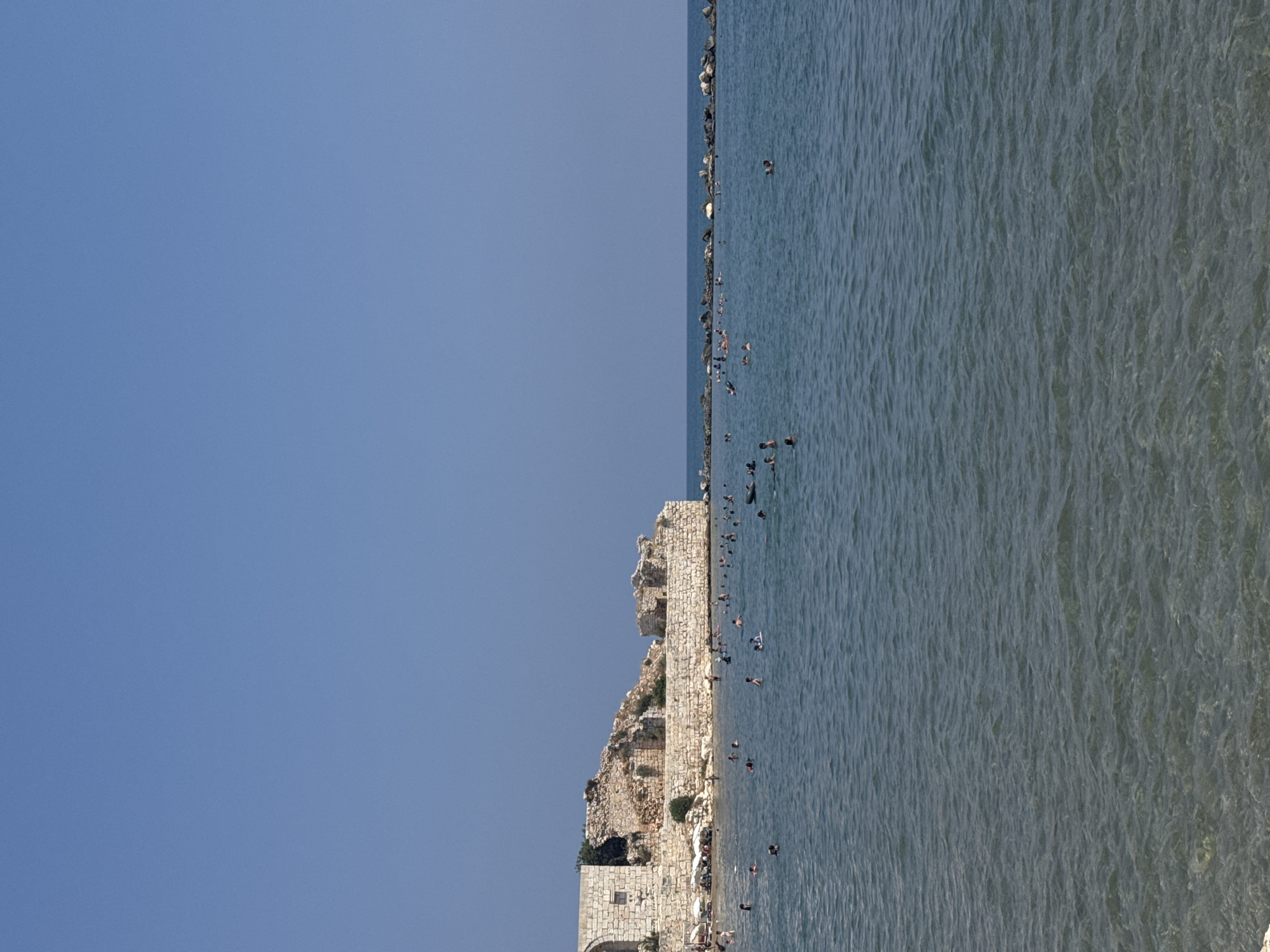 The image depicts a coastal scene in Erdemli, Turkey, featuring the ruins of a historical structure on the left, likely part of a castle or ancient building, constructed from large, weathered stone blocks. The ruins are situated along the shoreline, with some sections appearing intact while others are crumbled. In front of the ruins and extending into the sea is a beach area with numerous beach umbrellas. The sea itself is a calm, clear blue, with a significant number of people swimming, wading, and relaxing in the water. The horizon is marked by a clear, bright blue sky with no clouds. A breakwater or a line of large rocks extends from the right side of the frame into the sea, with more people visible near it. The water is shallow enough in some areas to see the seabed, which appears to be sandy or pebbly. The overall atmosphere is one of leisure and recreation on a sunny day at the coast.