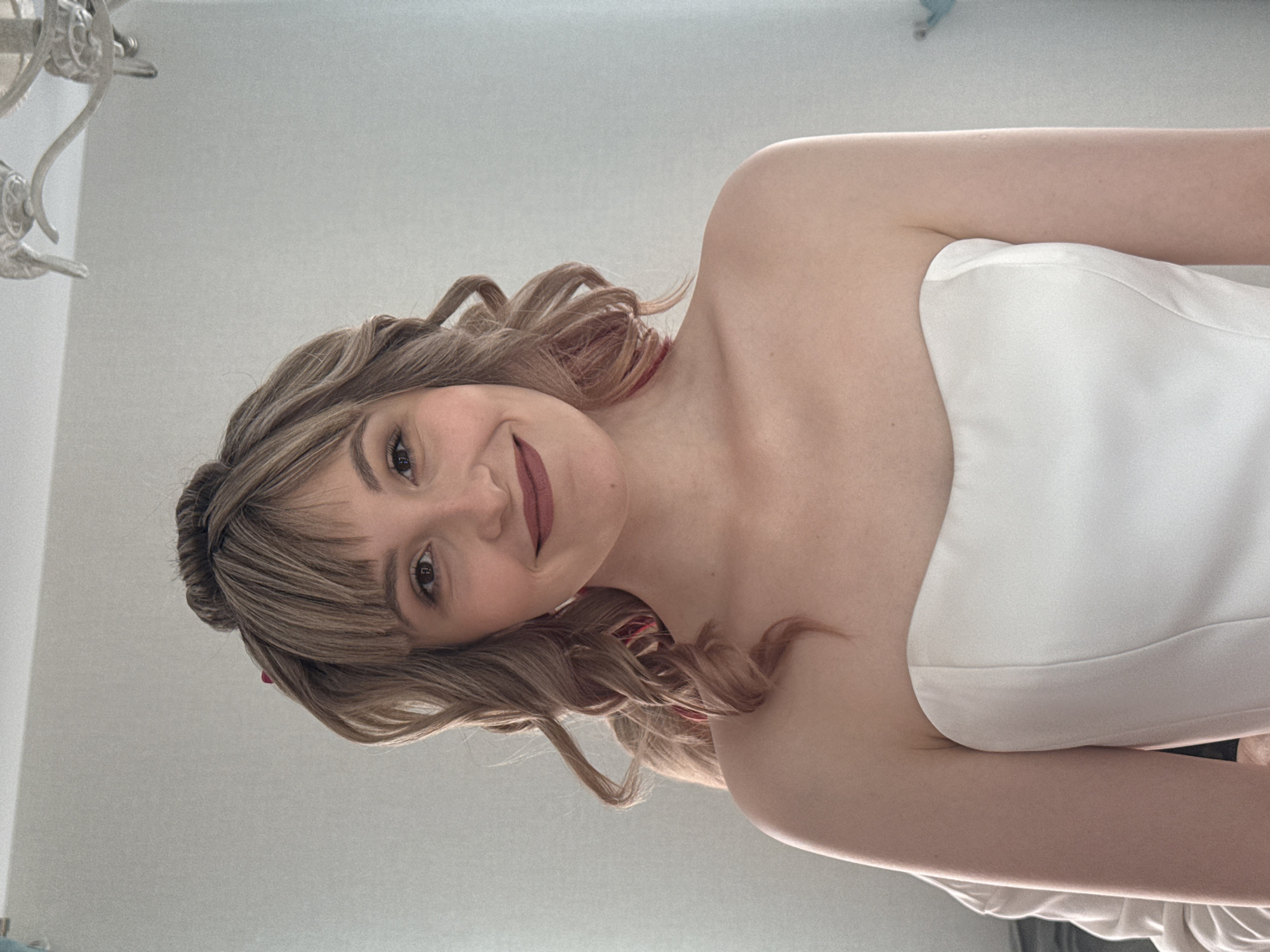 A young adult woman is captured in a close-up indoor portrait, taken from a low-angle perspective as she lies down, looking directly up at the camera with a warm smile. She has wavy, medium-length light brown hair styled with bangs across her forehead, and her hair appears to have subtle reddish highlights or strands. She wears a strapless white top or dress. Her makeup includes dark eyeliner accentuating her eyes and a matte, rose-toned lipstick. A hint of red fabric or an accessory is visible behind her neck.
The background consists of a plain, light-colored wall, likely white or light gray, suggesting a simple, clean indoor setting. In the upper left corner, a portion of an ornate, possibly metal, light fixture or ceiling decoration is visible. The lighting is soft and even, indicating bright indoor conditions. There is no visible text in the image. The woman's expression conveys happiness and contentment. While no specific architectural or outdoor cues are present, the provided context places this scene in Izhevsk, Russia.