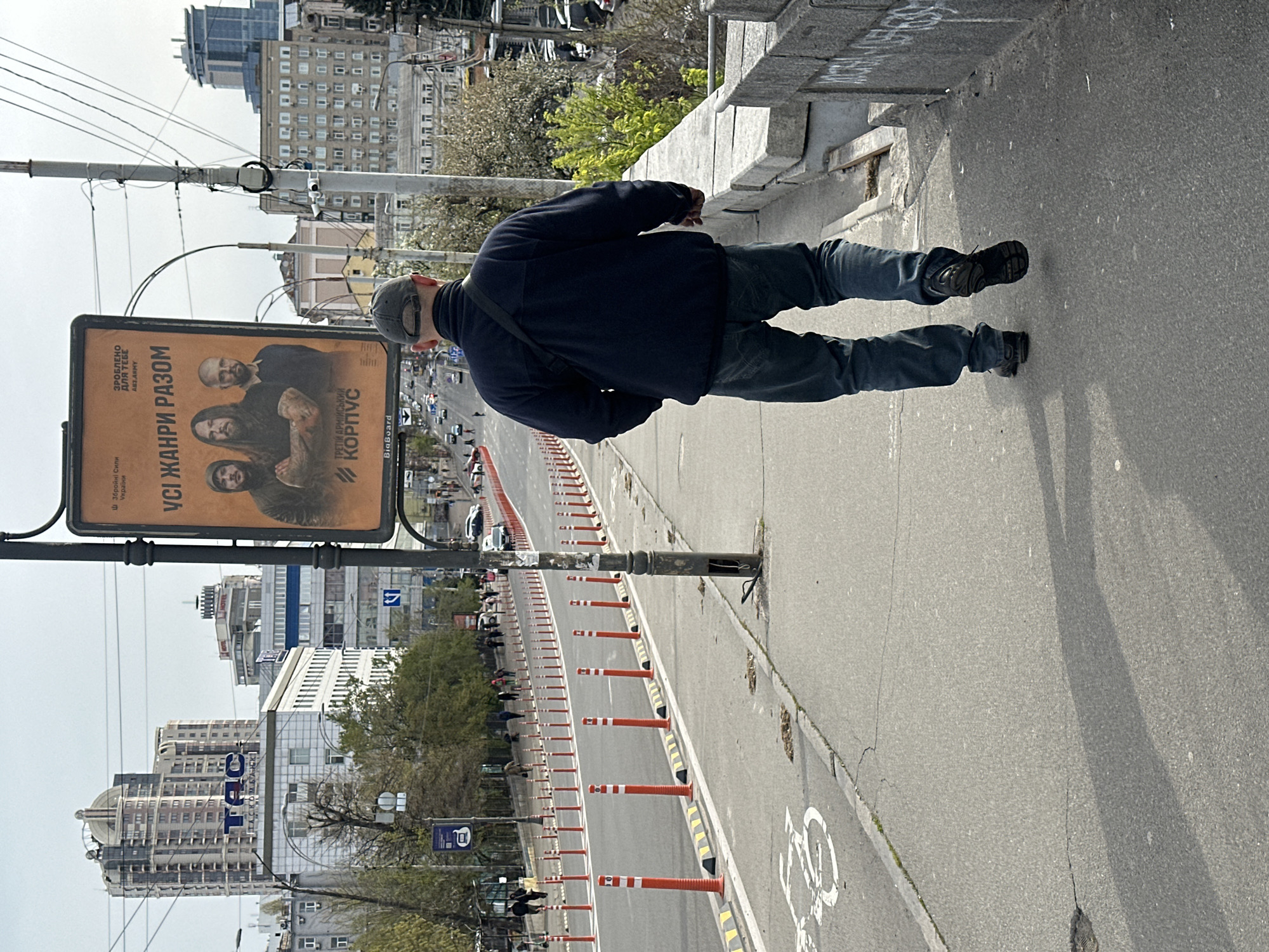 The image shows an urban scene in Kyiv, Ukraine. A man in a dark jacket and cap stands on a sidewalk near a bridge or overpass. Beside him is a large billboard featuring the text "УСІ ЖАНРИ РАЗОМ" (All genres together) and "ТРЕТІЙ АРМІЙСЬКИЙ КОРПУС" (Third Army Corps). The billboard includes the smaller text "ЗРОБЛЕНО ДЛЯ ТЕБЕ ABT ARMY" (Made for you AVT Army) and "Ш Збройні Сили України" (Armed Forces of Ukraine). Beyond the man and the billboard is a road with orange and white traffic cones. In the background, city buildings and trees are visible under a cloudy sky. One building features the letters "TAC". There are vehicles on the road. The sidewalk has a designated bicycle lane.