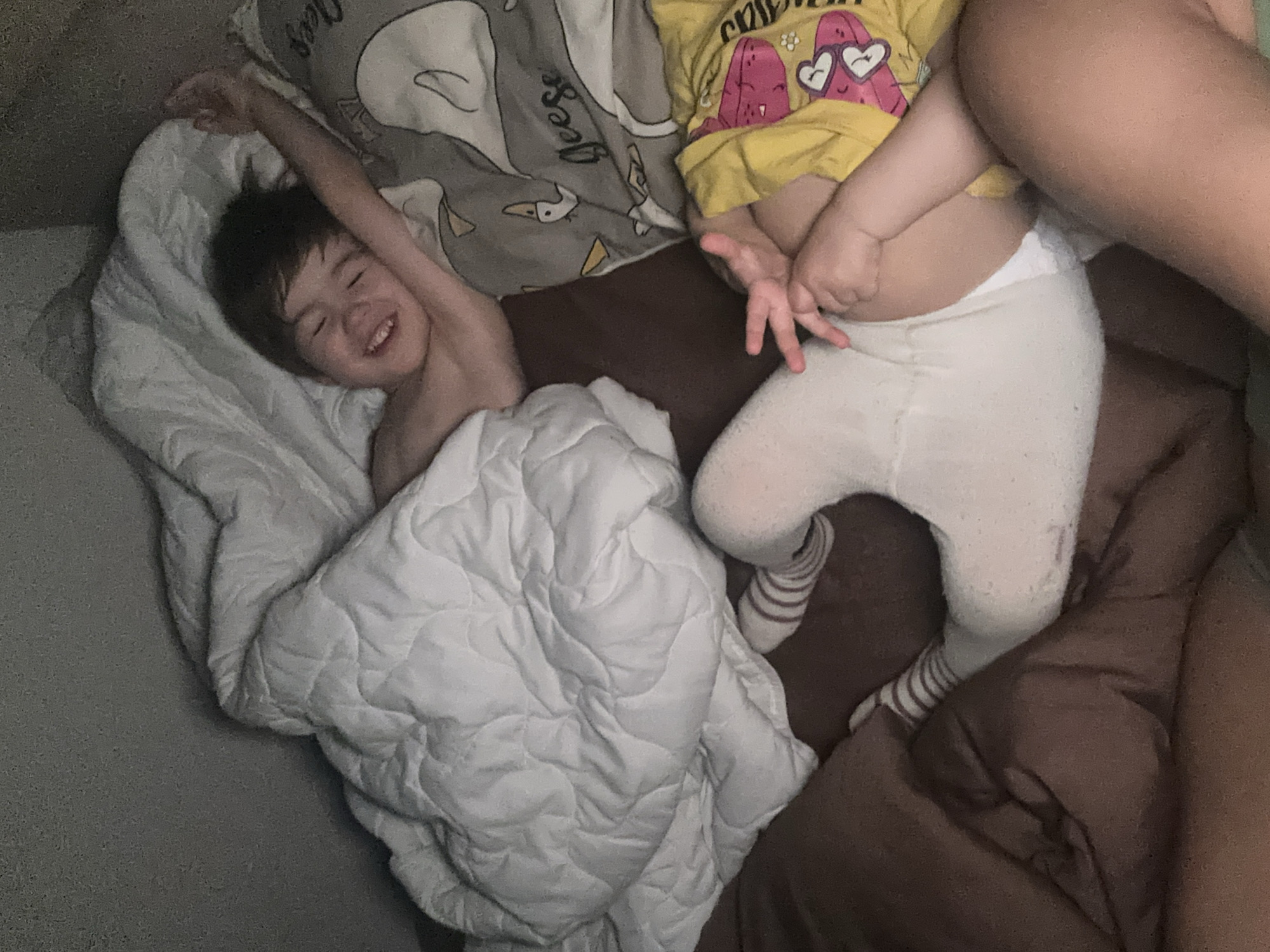 A young child is lying in bed, covered with a white comforter. They are smiling and have one arm raised in the air. Next to them is another child wearing a yellow shirt, white tights and socks. They are also lying on the bed, but their body is partially obscured by an adult. The background includes pillows with a cartoon design. The location is unknown.