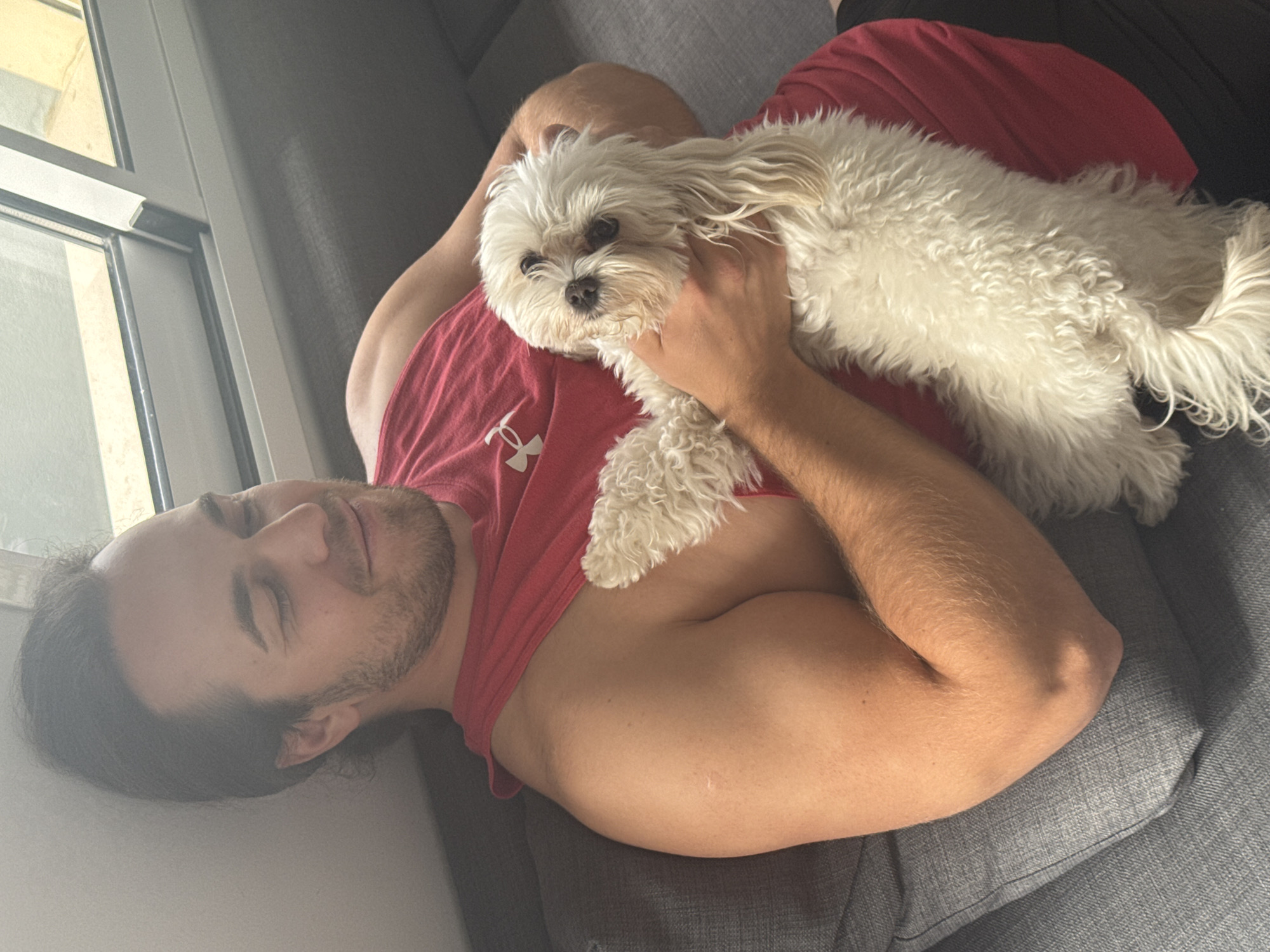 This image captures a cozy indoor scene featuring a man and a small white dog. The man is lying on his back on a grey upholstered couch, appearing relaxed or asleep with his eyes partially closed. He has dark hair and a beard, and is wearing a red t-shirt with a white Under Armour logo visible on his left chest. His arm, showing some musculature, is bent and resting near his side.
A small, fluffy white dog, possibly a Maltese or similar breed, is comfortably nestled on the man's chest. The dog has dark eyes and long, shaggy fur, and is looking directly towards the camera. The interaction suggests a moment of close companionship and affection.
To the left of the man, a window with light-colored framing is visible, allowing bright daylight into the room, indicating it is daytime. The background through the window is indistinct but suggests an exterior building or wall. A light-colored wall is also visible behind the man's head. The overall atmosphere conveys a sense of peacefulness and comfort. This scene takes place in Valencia, Spain.