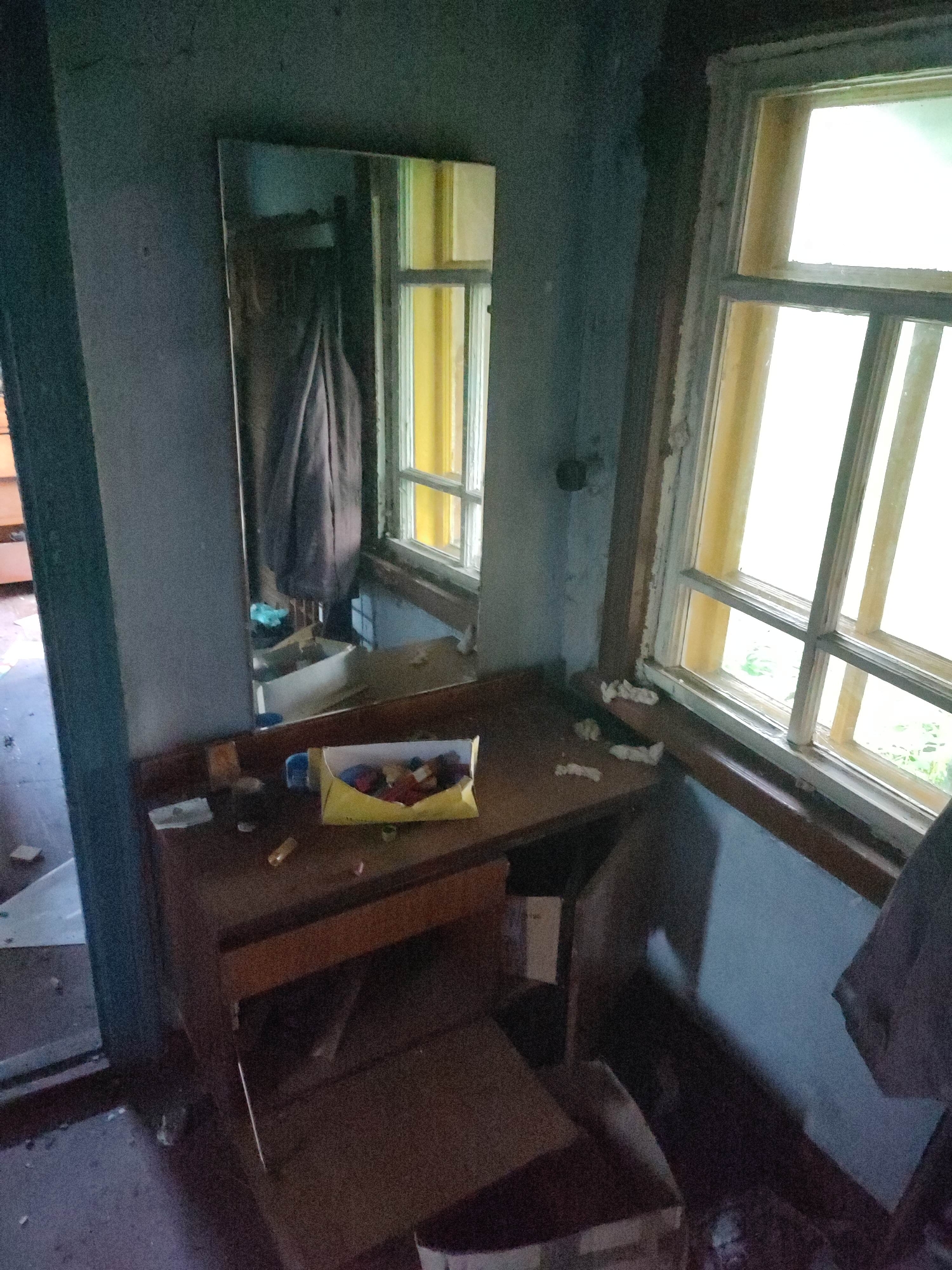 This image depicts an interior scene, likely an abandoned or neglected room, within the city of Gremyachinsk, Russia. The room contains a wooden vanity table with a mirror. On the vanity, there is a yellow box filled with small colorful items, possibly craft supplies or buttons, along with other assorted objects like a tube and some paper scraps. A drawer is partially open in the vanity. To the right of the vanity, a window with a wooden frame is visible, showing daylight outside. The walls are a dark, muted color, and the floor appears dusty and littered with debris. A dark fabric, possibly clothing, is hanging on the wall to the left of the mirror. The overall impression is one of disuse and decay.