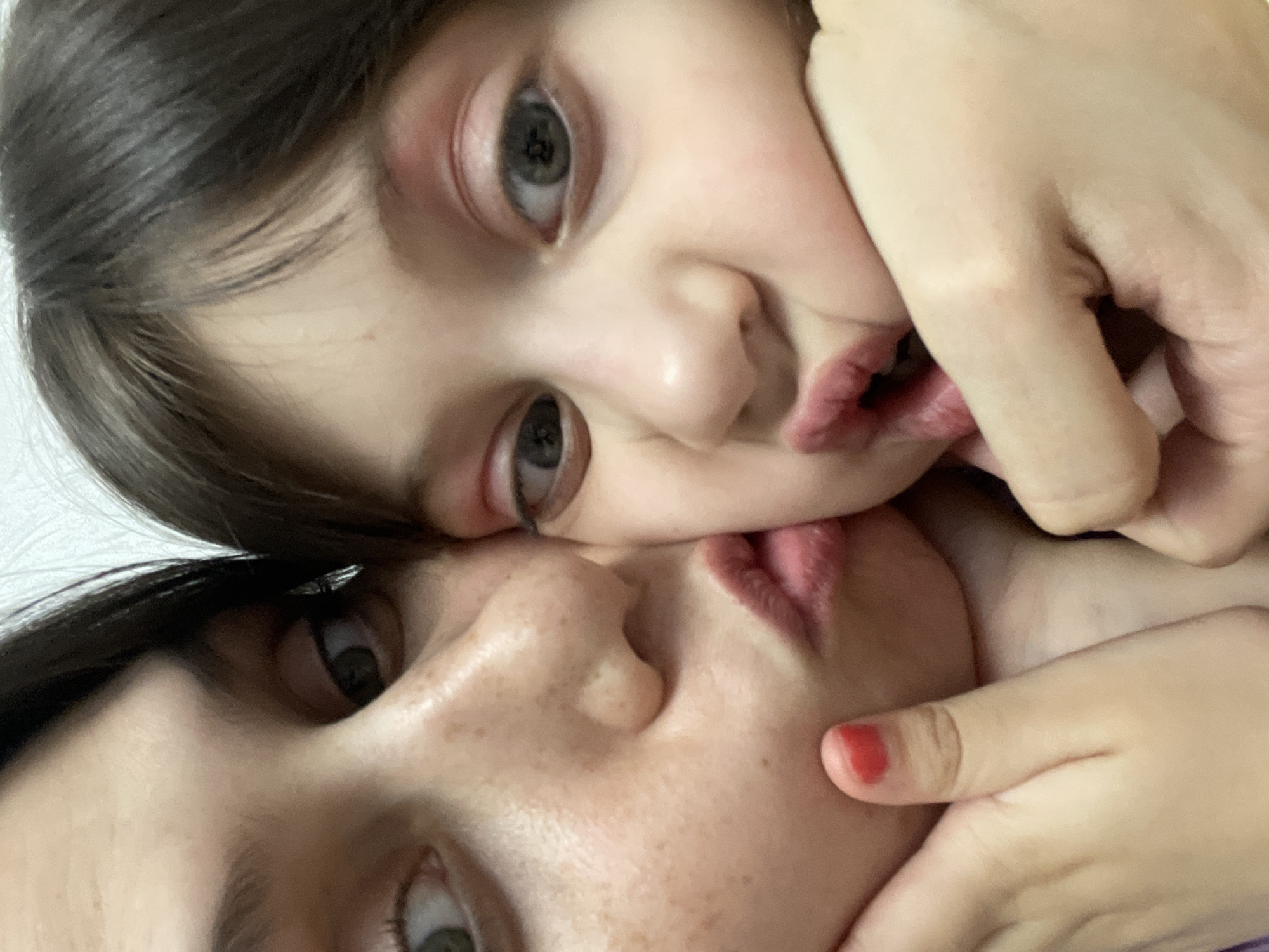 The image is a highly-rotated, close-up portrait featuring two individuals, likely a mother and child, engaged in an affectionate interaction. The two faces are positioned very intimately, with their noses almost touching and lips slightly puckered, suggesting a kiss or a playful pose.
The subject in the upper part of the frame, presumed to be the child, has dark brown hair, fair skin, and striking, wide-open eyes with light irises that look directly at the viewer. Their small hand gently rests on the cheek of the other person. The lower subject, likely the adult, has dark hair, visible freckles across their nose and cheek, and eyes open but angled slightly away. A hand belonging to the adult, with a finger painted with red nail polish, is visible near their mouth, possibly cradling the child's face or their own.
The close proximity and gentle contact of their faces and hands emphasize a deep emotional connection and tenderness. The background is soft, out of focus, and appears to be a plain, light-colored surface, suggesting an indoor setting. The lighting is soft and even, illuminating their features. The overall impression is one of love, warmth, and intimacy.
No text is visible in the image. The provided location context is Izhevsk, Russia.