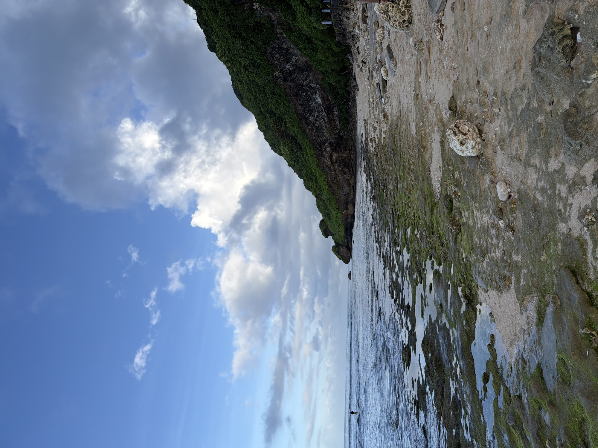 The image depicts a coastal scene at low tide in Kuta Selatan, Indonesia. A rocky shoreline with algae-covered stones stretches along the bottom of the frame, meeting the ocean's edge. Further up, a cliff covered in dense green vegetation dominates the upper right, while the sky occupies the left side, filled with blue patches and scattered white clouds. The horizon appears to be at eye level.