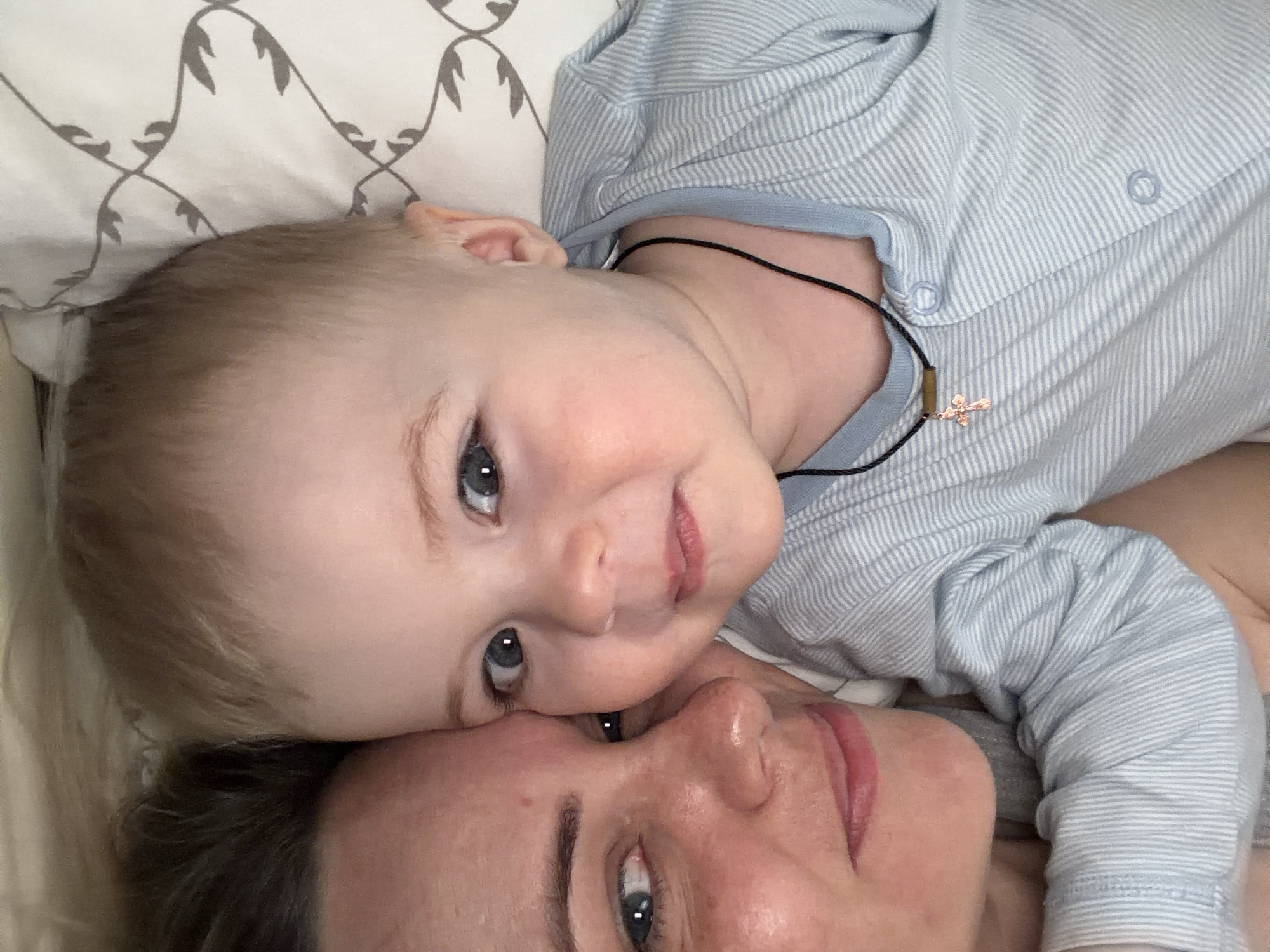 A close-up, overhead shot shows a baby and an adult lying on what appears to be a bed. The baby is fair-skinned with blue eyes and light blonde hair. The baby is wearing a light blue striped onesie and a black necklace with a small gold-colored cross pendant. The baby's head rests on the adult's face. The adult has fair skin and dark hair. The background is a light-colored pillow with a patterned design. There is no indication of the location being Votkinsk, Russia.
