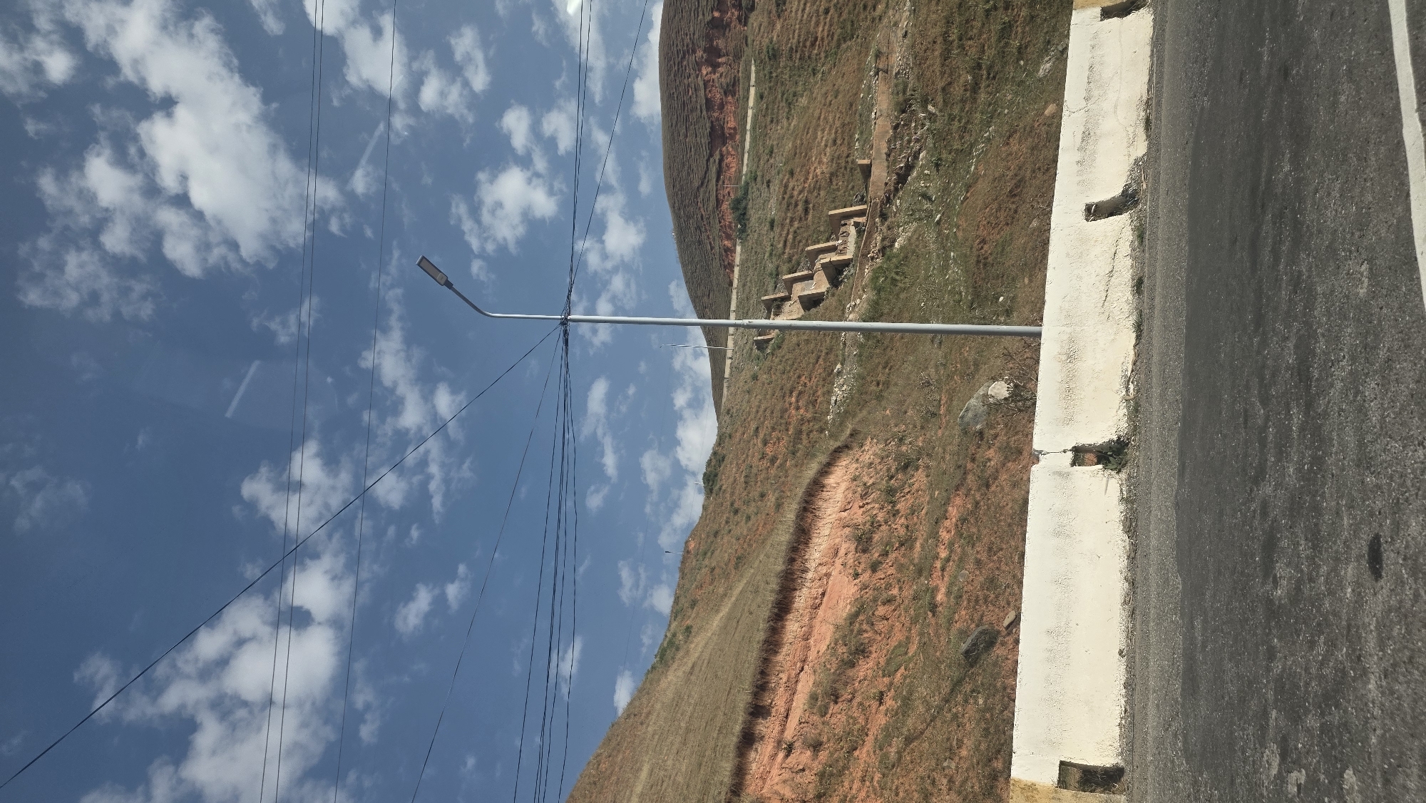 The image shows a hillside with sparse vegetation and exposed reddish-brown soil. A concrete barrier runs along the foreground, separating a road from the hillside. A tall streetlamp stands on the hillside, and power lines stretch across the sky. In the distance, there are visible remnants of what appear to be old stone structures or a staircase leading up the hill. The sky is blue with scattered white clouds. The scene is devoid of any people. The image was taken on a sunny day. The location is identified as being in the city of Taldyk, Kyrgyzstan.