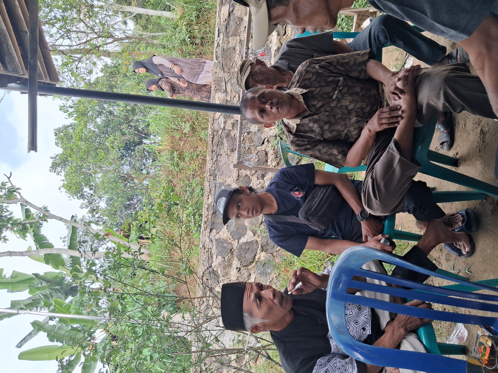 In Musuk, Indonesia, a group of men are gathered outdoors, seated on plastic chairs around a low table. The environment is rural, with lush green foliage, banana trees, and a stone wall in the background, suggesting a hillside location. The sky is overcast.
The central figures are four men engaged in conversation. The man on the far left, wearing a black peci (a cap common in Indonesia) and a black shirt, is smoking a cigarette. Next to him, a younger man wears a black t-shirt and a black baseball cap. To their right, a man in a patterned brown shirt is speaking, with his hands resting on his knees. Behind him, another man wears a cap and a grey shirt. Further in the background, two women in headscarves are visible. The overall atmosphere appears relaxed and informal, with the men in conversation and casual attire.