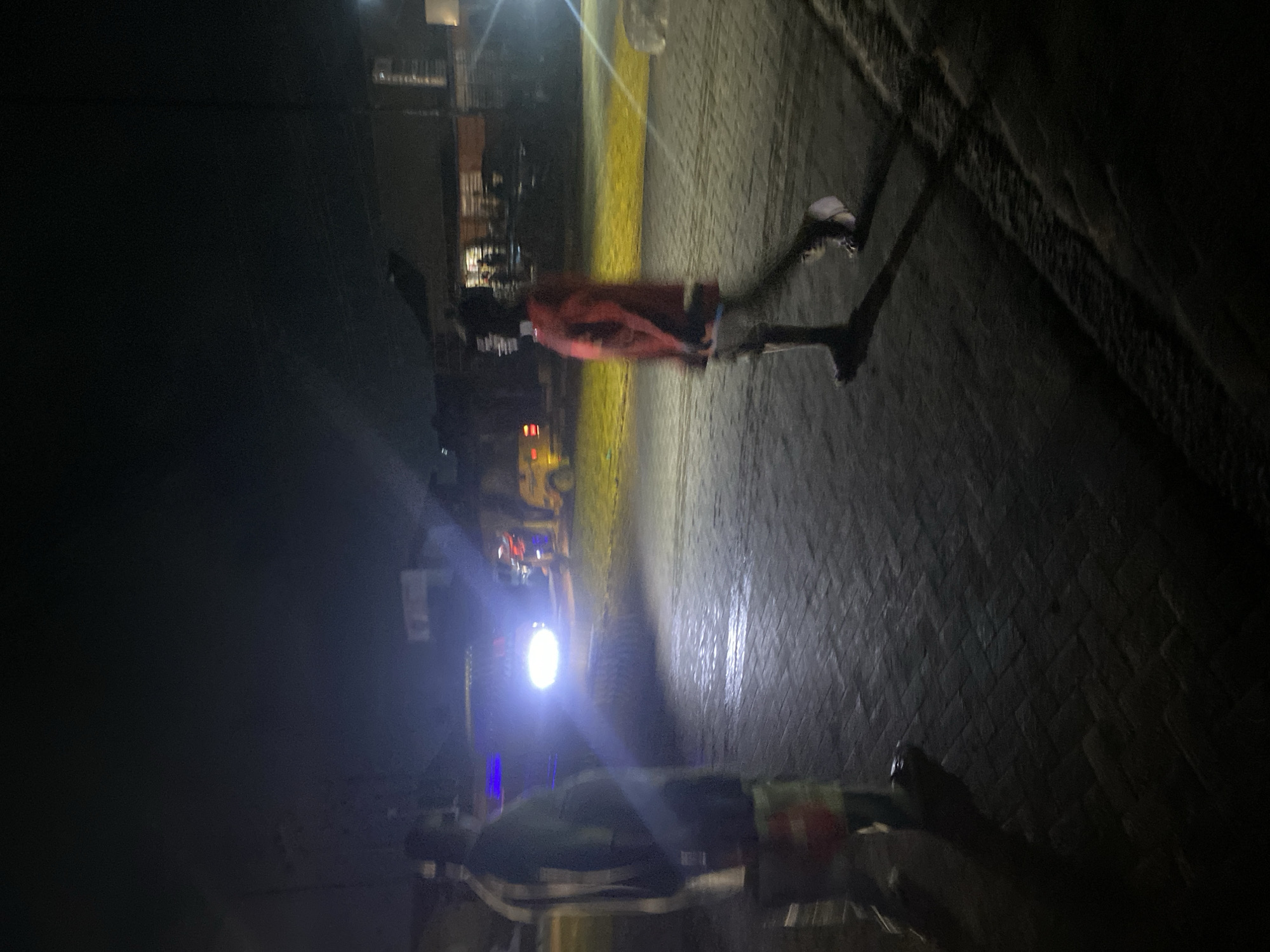 This image depicts a nighttime scene on a paved roadway or courtyard in Ikorodu, Nigeria. The ground surface is composed of dark, interlocking pavers, exhibiting reflections from various light sources, indicating a wet surface or high reflectivity.
In the left mid-ground, a person is observed from behind, facing right. This individual wears a dark garment, possibly a uniform, with a discernible orange or red reflective patch on the lower back or hip area. Their form exhibits slight motion blur, consistent with movement or a long exposure.
In the right mid-ground, a second person is observed from the left side, walking towards the left. This individual wears a red long-sleeved top and dark lower garments. Significant motion blur is evident in their legs and arms, confirming active locomotion. A pronounced, elongated shadow is cast by this individual onto the paved surface.
In the central background, a large vehicle, possibly a truck, is positioned with its bright headlights actively illuminated, casting a strong beam onto the pavement. Behind this vehicle, faint blue emergency lights are visible, and further to the right, a yellow vehicle is partially discernable in the background. Distant buildings and structures are visible in the far background under low illumination. The sky is dark, indicating a complete absence of natural light. Multiple artificial light sources contribute to areas of high illumination and deep shadows throughout the scene.