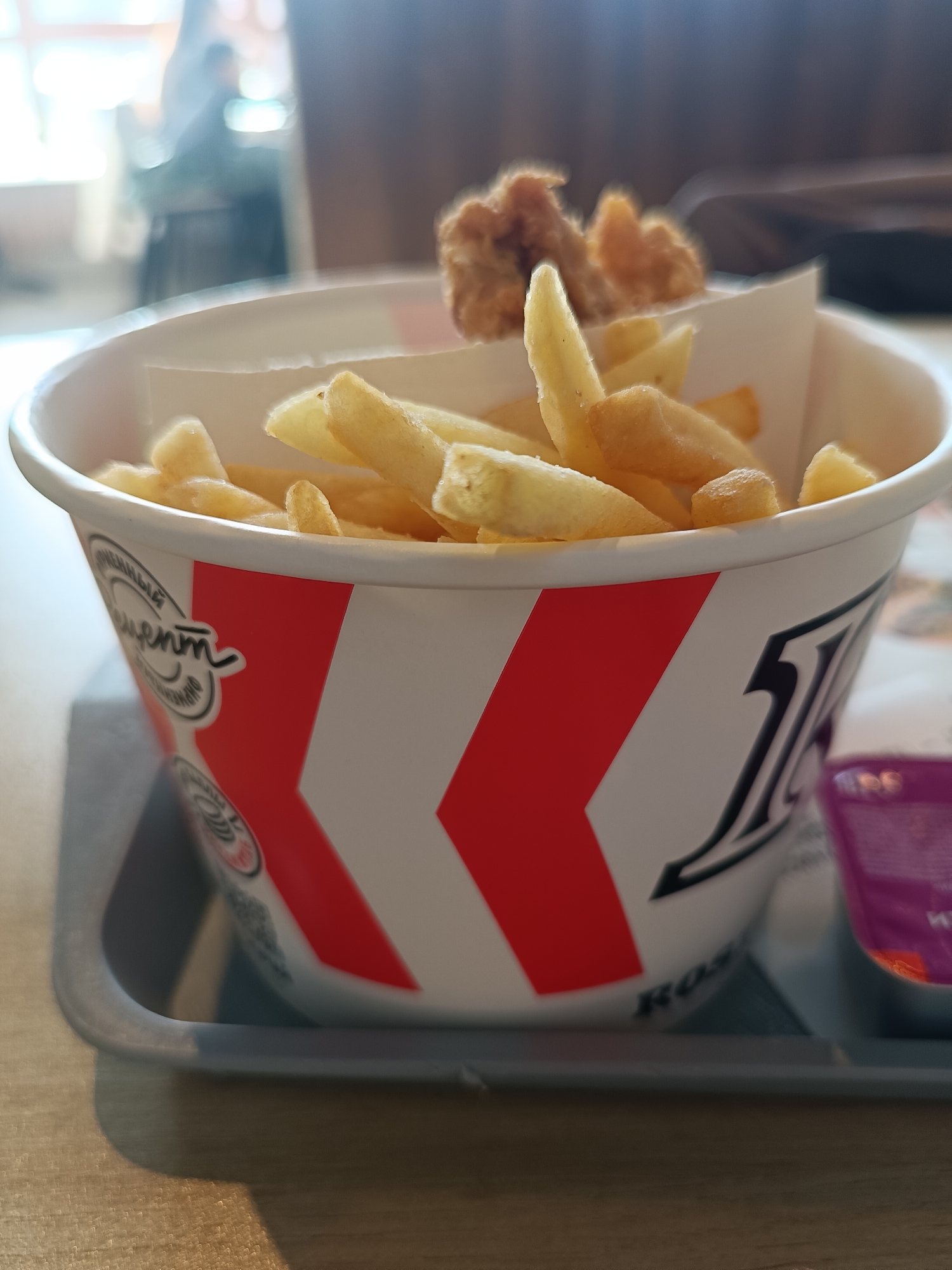 A medium shot captures a paper bucket filled with french fries and a couple of chicken nuggets, placed on a grey tray. The bucket is decorated with white and red chevron patterns and appears to be from a fast-food restaurant. The tray also contains a small container of sauce. The location appears to be a fast-food restaurant.