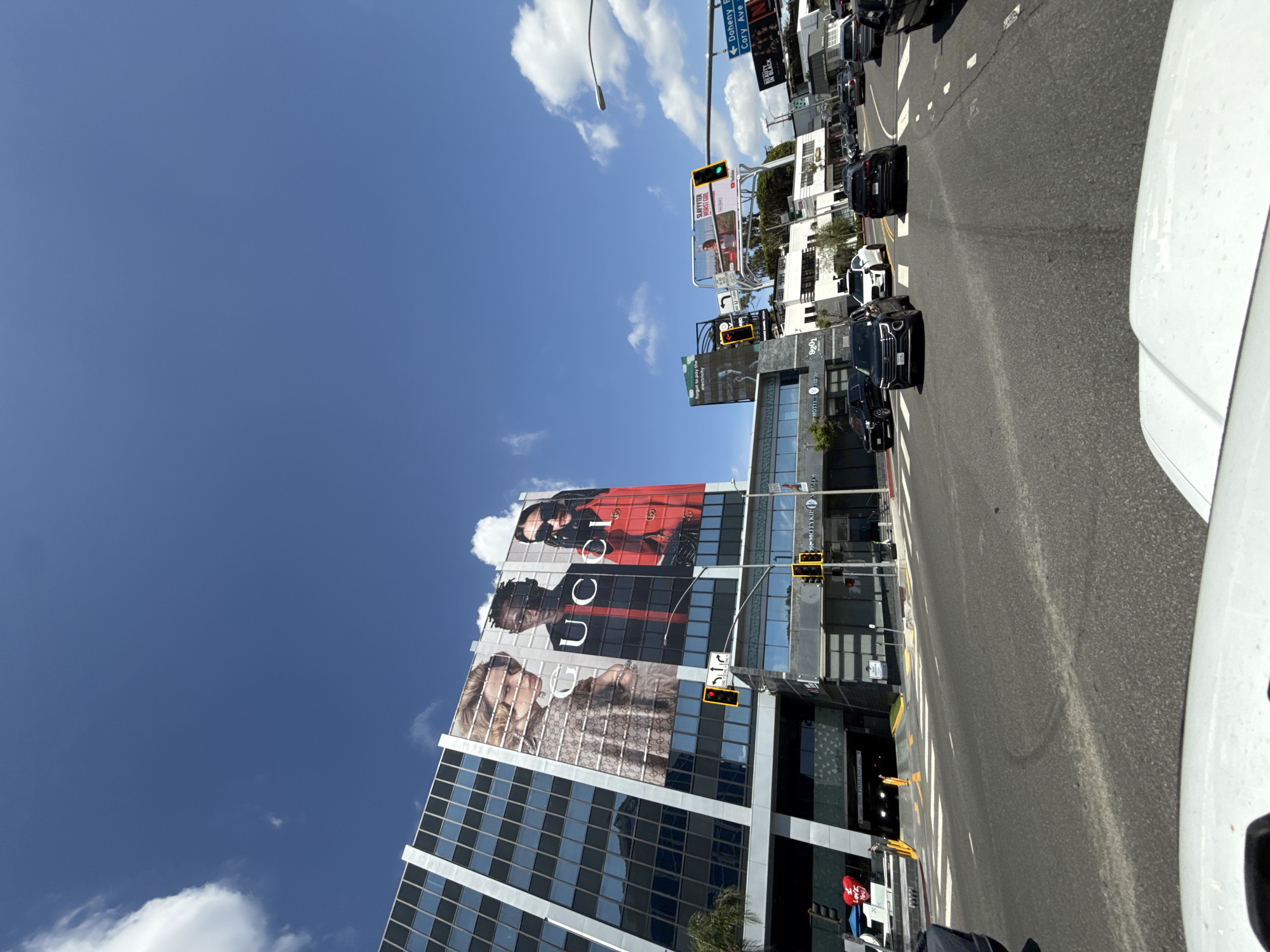 The image captures a street scene in West Hollywood, California, featuring a modern building with a large Gucci advertisement on its side. The building is a multi-story structure with a grid-like pattern of windows, and the ad showcases images of people wearing Gucci clothing.
Traffic lights are visible along the street, and several cars are driving on the road. There are also multiple billboards of advertisements including beauty in Black and the YouTube streamer slaysslayter. Street signs are visible, including signs directing toward "Doheny" and "Cory Ave." "Motykiswede" is also advertised.
The sky is bright blue with scattered clouds. The image appears to have been taken from the perspective of someone inside a vehicle on the road.