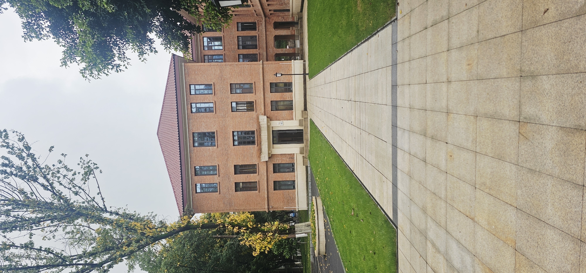 The image shows a three-story brick building with a red tiled roof, likely located in Yuzhong District, China. The building has multiple windows and a central entrance. A paved walkway leads towards the building, flanked by strips of green grass. Trees with green and yellow foliage are visible on both sides of the frame. The sky is overcast.