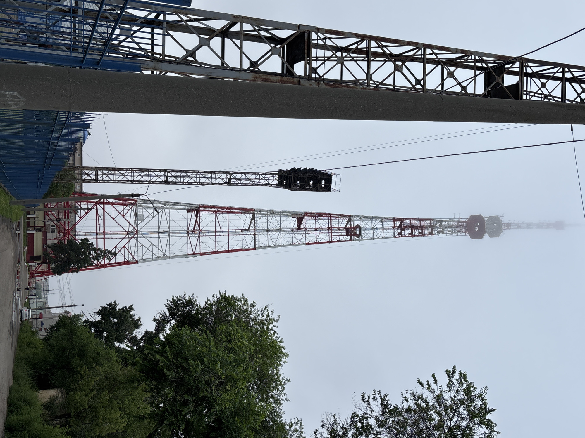 This image shows a view of a telecommunications tower in Blagoveshchensk, Russia, on an overcast day. The primary subject is a tall, red and white lattice tower, partially obscured by fog at its upper reaches. To the left of the main tower, a smaller, darker lattice structure with what appears to be lighting equipment is visible. In the foreground, a concrete utility pole stands prominently, with a weathered, rust-colored lattice structure to its left and a blue metal fence running along the side. Trees with lush green foliage are present on the right side of the image, framing the scene and partially obscuring buildings in the background. A dirt road leads towards the telecommunications tower, with a white car parked on the right side of the road and a few people walking in the distance. The environment appears to be an industrial or semi-industrial area, with signs of infrastructure and some minor water puddles on the ground.