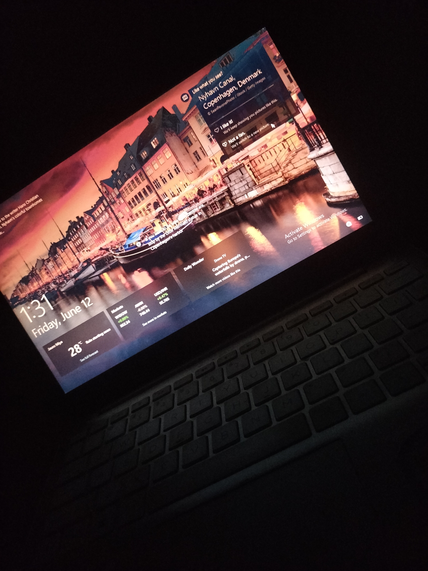 The image shows a laptop with its screen illuminated, displaying a Windows lock screen. The lock screen features a scenic image of Nyhavn Canal in Copenhagen, Denmark, with colorful buildings lining the waterfront and boats in the canal. The time displayed is 1:31, and the date is Friday, June 12. There is also a weather icon showing 28 degrees Celsius and a forecast indicating rain starting soon. Additional information on the screen includes stock market data and a prompt to "Activate Windows." The laptop is placed on a dark surface, possibly a desk, and part of the keyboard is visible. The overall lighting is dim, suggesting the image was taken in a low-light environment. There are no people visible in the image. The location in the city of Jalingo, Nigeria, is not depicted or implied in this image.