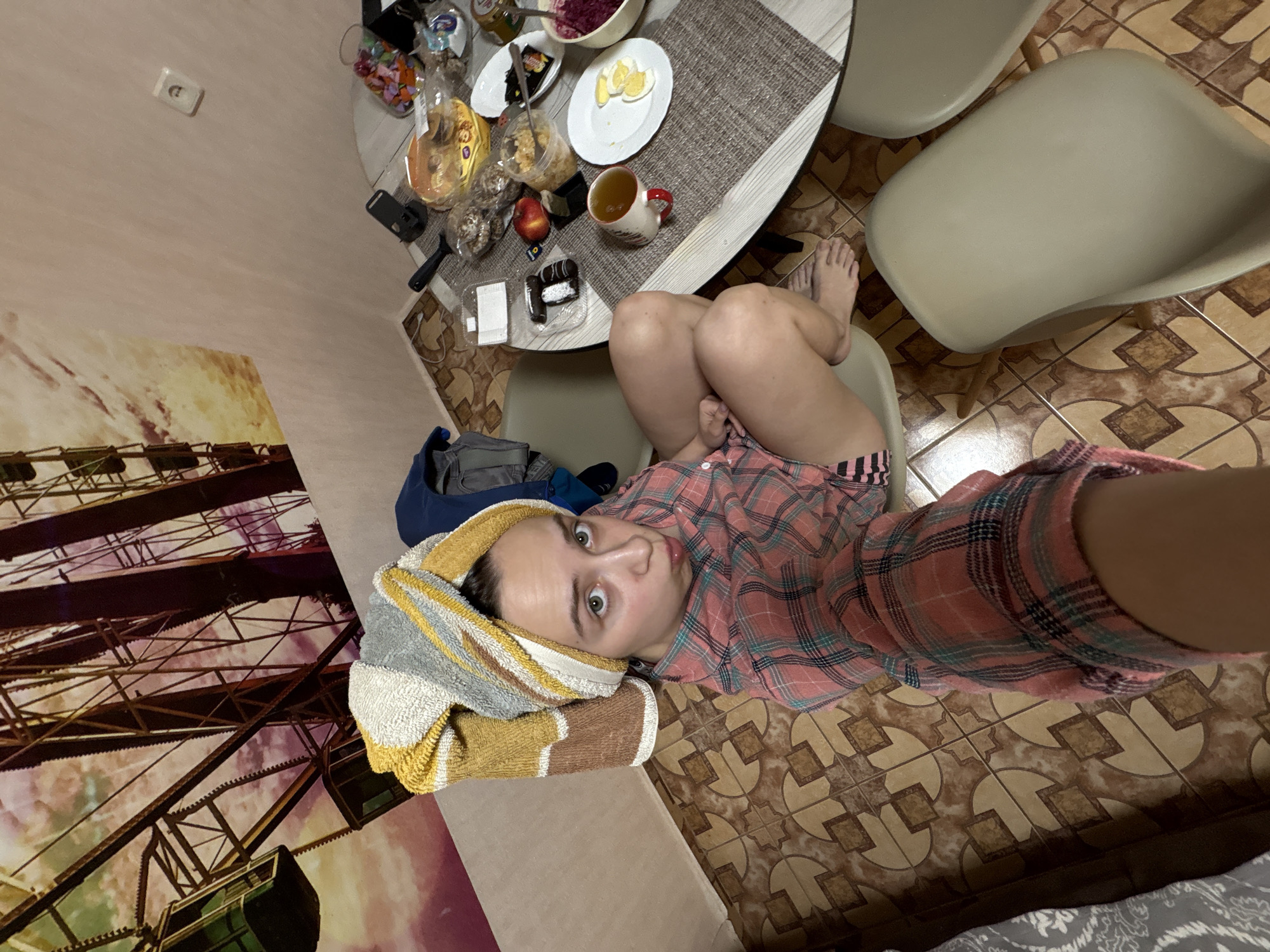 An indoor scene, likely a kitchen or dining area in a home in Прикамский, Russia, is captured from a high, tilted angle. The primary subject is a person, appearing female, seated at a round wooden table. She looks directly at the camera with wide, slightly surprised eyes and a playful expression, her lips slightly pursed. She wears a pink plaid shirt and has a striped towel (yellow, grey, and white) wrapped around her head, suggesting she may have recently showered or washed her hair. Her legs are visible, crossed, as she sits on a modern light-colored chair with wooden legs.
The table is covered with a striped placemat and holds various food items. These include a bowl of what appears to be beetroot salad, a plate with sliced boiled eggs, a white mug with a red handle containing a beverage, a red apple, and several packaged snacks like pastries, donuts, and candies in clear containers. A black spatula and a smartphone in a stand are also on the table. Another similar empty chair is visible nearby.
The floor is tiled with a brown and light beige geometric pattern. The wall is a light, neutral color, with a small white light switch or outlet. A large wall print or poster depicts a Ferris wheel against a sky with warm and cool hues, possibly suggesting sunset or dusk. A blue bag or backpack is partially visible on the floor near the person. No legible text is visible in the image.