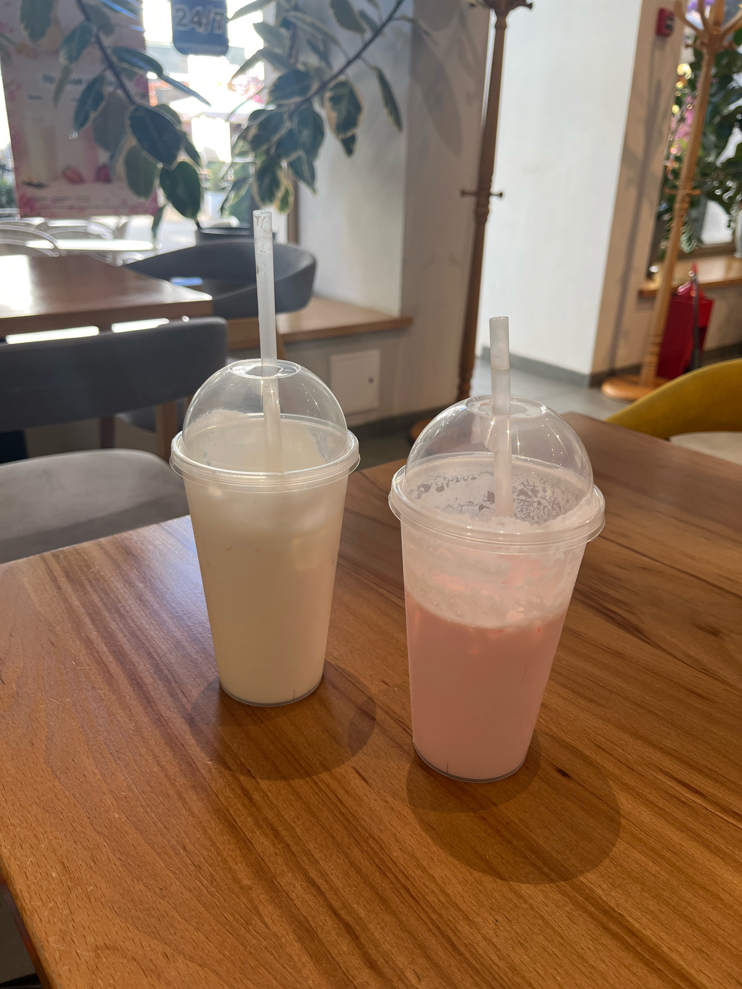 Two plastic cups with clear dome lids and white straws sit on a wooden table. The cup on the left contains a white beverage, and the cup on the right contains a pink beverage. The table has a wood grain pattern. In the background, there are chairs, tables, and plants, suggesting an indoor cafe or restaurant setting. The lighting indicates it is daytime, with sunlight streaming in from a window. The scene appears to be in Simferopol, Ukraine.