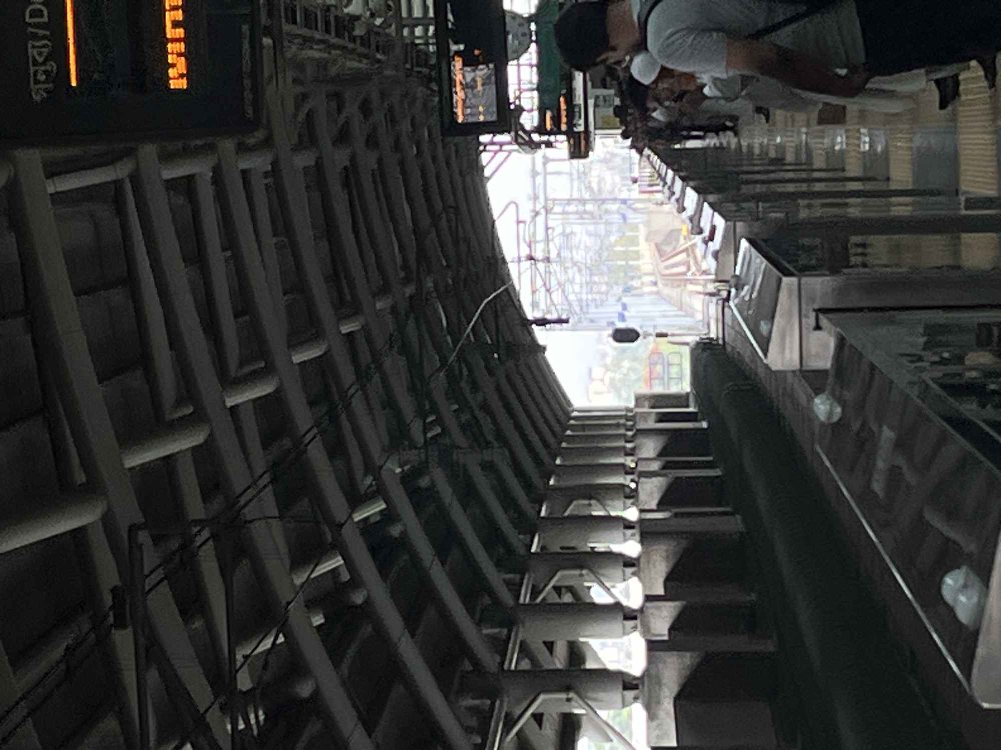 The image shows an interior perspective of a Dhaka Metro Rail station, Bangladesh. Passengers are lined up at ticket counters. Overhead is a structural framework of steel beams and cables. A digital display screen is visible, along with other station signage written in Bengali. The daylight coming from the station exit illuminates the inside.