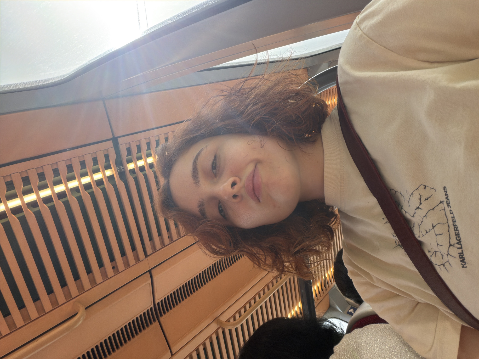 A young woman with reddish-brown, curly hair is taking a selfie inside a vehicle. She is wearing a light-colored t-shirt with a Karl Lagerfeld Jeans graphic. A dark red strap is visible across her shoulder. The interior of the vehicle features a tan ceiling with parallel slats and lighting fixtures. A window with bright light shining through is to her right. To her left, another person with dark hair is partially visible. The setting appears to be a public transport vehicle, possibly a train or bus. There is no specific indication of the location being Strasbourg, France.