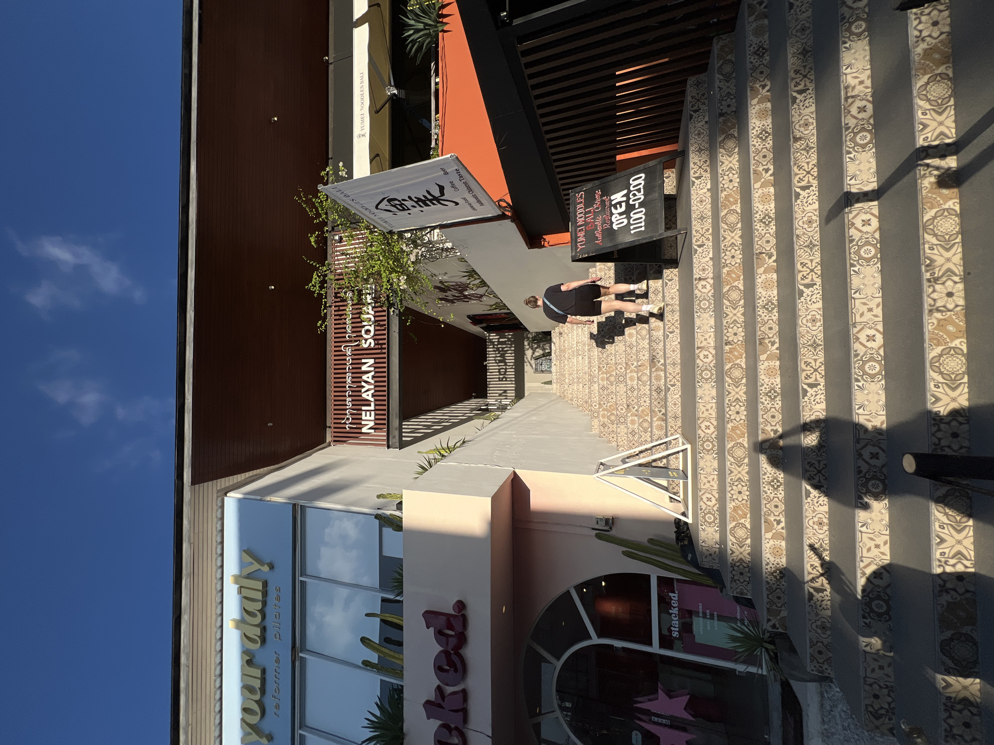 A street scene in North Kuta, Indonesia, features stairs leading up to "Yumei Noodles Bali," indicated by a signboard stating "Open 1100-02.00." A woman is walking up the patterned stairs. Other establishments are visible, including "your daily reformer pilates" and "stacked," a restaurant or bar. "Nelayan Square" is also displayed. The sky is clear and blue.