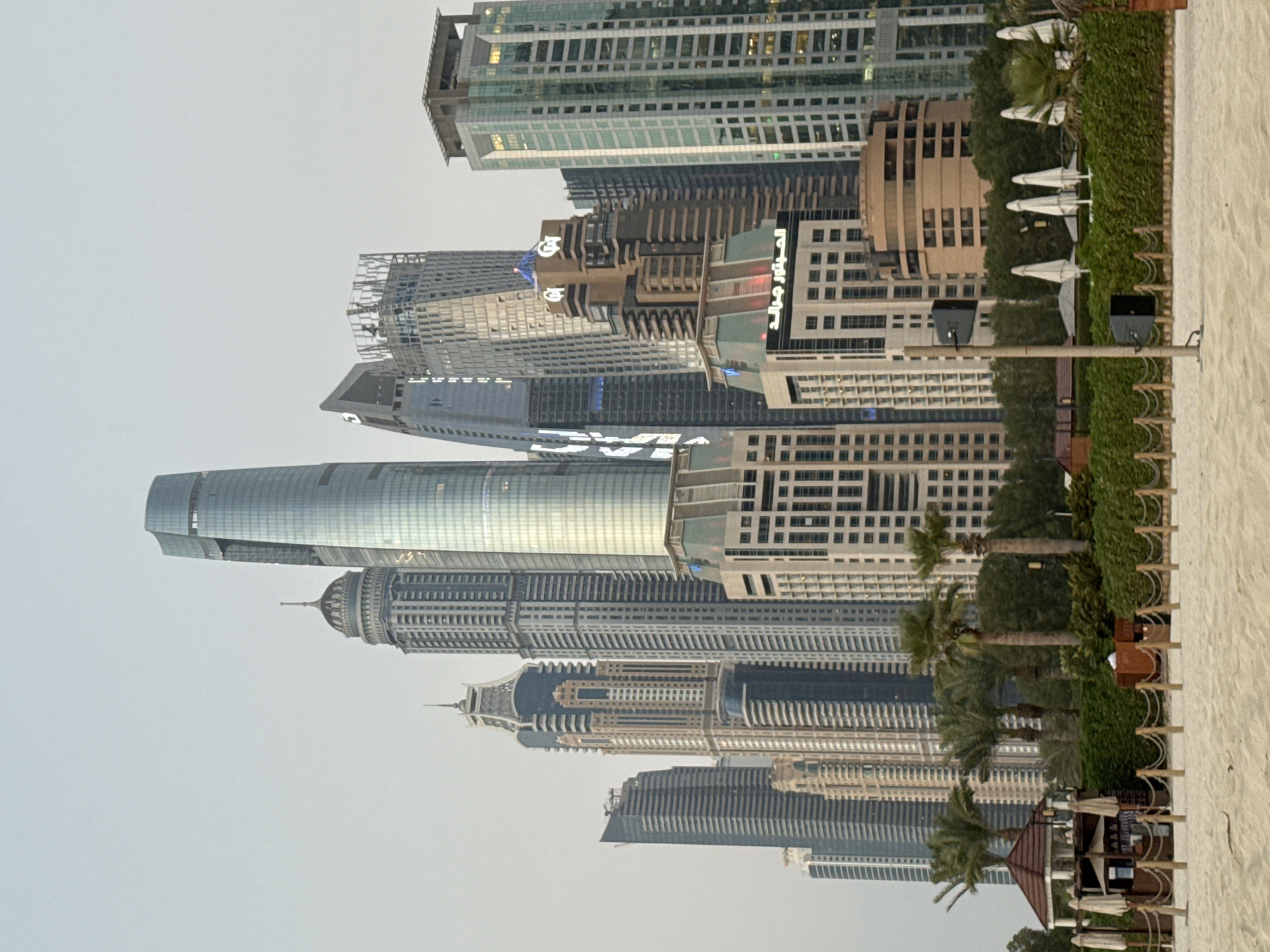 This image depicts a skyline of modern skyscrapers in Dubai, United Arab Emirates. The buildings are a mix of glass and concrete, with varying architectural styles, including some with curved facades and others with sharp, angular designs. Some buildings appear to be under construction, indicated by scaffolding. In the foreground, to the right, a sandy beach is visible, with several white beach umbrellas and palm trees suggesting a resort or public beach area. The sky is overcast and pale, suggesting either early morning or late afternoon. The overall scene is one of urban development and leisure. No people are clearly visible in the image. There is some Arabic text on a building facade, which appears to be "المطور جالد" or similar, meaning "Developer Galad".