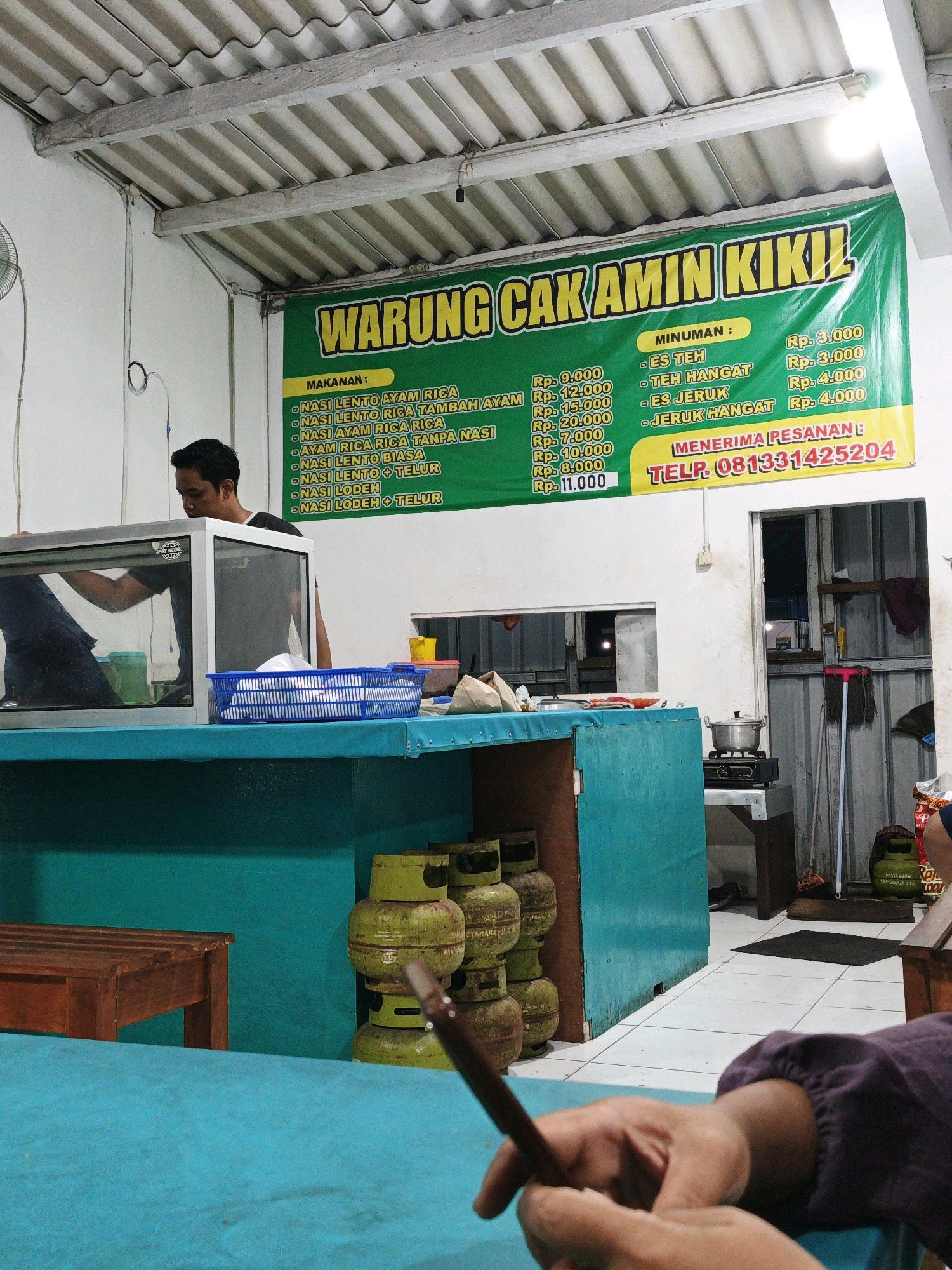 The image depicts the interior of "Warung Cak Amin Kikil," a food stall located in Kepadangan, Indonesia. A large green banner with yellow lettering announces the establishment's name and lists food and drink prices. The banner also displays a phone number for orders.
In the foreground, a person's hands are visible, holding what appears to be a wooden chopstick or similar utensil, suggesting they are about to eat or are in the process of preparing food. The person's upper body is slightly out of focus, but they are wearing a dark purple long-sleeved shirt.
Behind the hands, a bright turquoise-covered surface, likely a table, dominates the lower portion of the frame. To the left, a wooden bench or seat is partially visible.
Further back, a glass display case with a silver frame contains food items. A man with short dark hair, wearing a dark t-shirt, is standing behind the display case, reaching into it.
The background reveals a simple interior with white walls and a corrugated metal ceiling. Stacked green gas cylinders are visible to the right, indicating cooking facilities. A small stove with a pot on it is also present. The overall environment appears to be a humble, local eatery.
