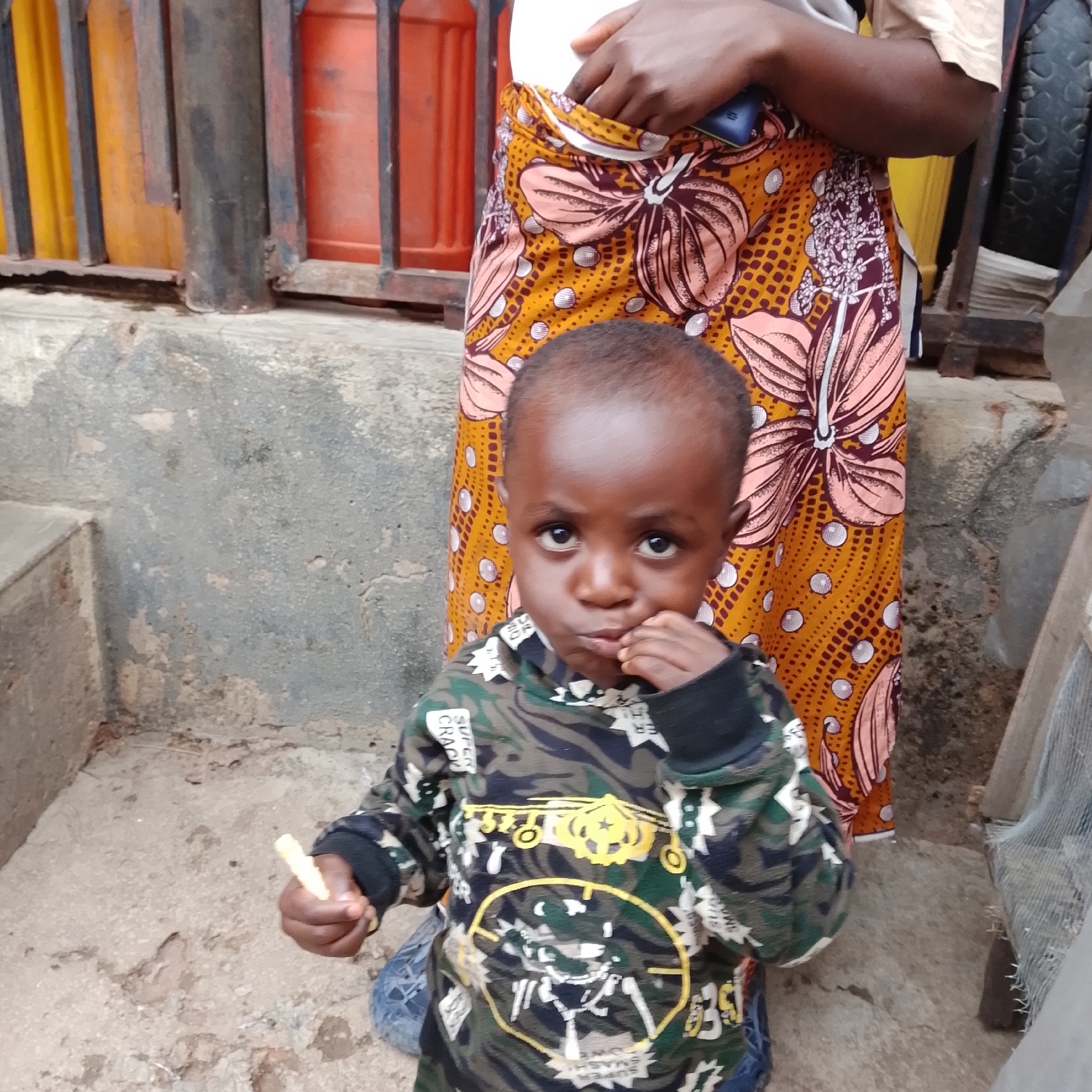 A young child with dark skin and short, dark hair is the primary subject of this medium-shot photograph. The child is wearing a camouflage-patterned long-sleeved shirt with yellow and white graphics, including what appears to be an airplane and a target symbol. The child is holding a small, light-colored object, possibly food, in their right hand and bringing it towards their mouth, suggesting they are eating. Their expression is somewhat serious, with eyes looking directly at the camera.
Behind the child, a person's lower body is visible, wearing a vibrant orange and yellow patterned fabric skirt with large floral designs in pink and white. The person's hand is partially visible, reaching into the folds of the skirt, possibly to retrieve something or adjust the garment.
The setting appears to be outdoors, with a concrete ground that is rough and textured, showing some cracks and discoloration. In the background, there is a dark metal railing or fence in front of vertical structures that are orange and yellow, possibly containers or doors. To the right, a rough, textured wall and some debris are visible, including what looks like a discarded tire. The lighting suggests it is daytime, but the specific time of day and weather conditions are not discernible. The overall atmosphere is candid and everyday, capturing a moment of the child's activity in an outdoor, possibly urban or semi-urban environment. No specific text is clearly legible on the child's clothing apart from some fragmented words or symbols that are part of the graphic design.