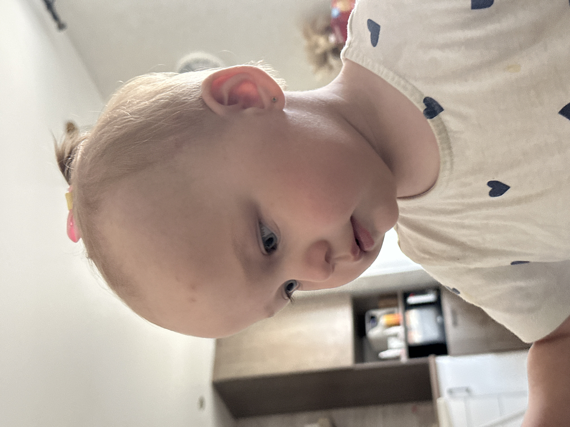 A close-up, eye-level, medium shot shows a fair-skinned Caucasian toddler with light blonde hair styled in a small ponytail and a pink bow, looking downwards and to the left. The toddler's face is illuminated by soft, natural light, highlighting their blue eyes, rosy cheeks, and parted lips. They are wearing a cream-colored t-shirt adorned with a pattern of small, dark blue hearts. An earring is visible in their right earlobe. The background is blurred, revealing hints of a kitchen interior with light-colored cabinets, countertops, and what appears to be a microwave and shelves with various items. The scene is set in a home environment, likely in the city of Chaikovsky, Russia.