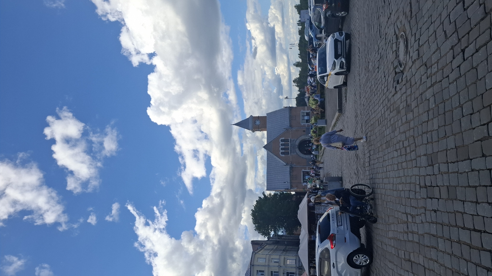 The image depicts a public square in Vyborg, Russia, on a cloudy day. In the foreground, a cobblestone pavement dominates the scene, with several parked cars visible on the right. A white sedan, identified as a Lada, is parked on the left.
Further back, a building with a distinctive clock tower stands prominently, appearing to be a historical structure, possibly a market or public administrative building. The architecture suggests it's a significant landmark in Vyborg.
Several individuals are present in the square. A woman in a blue and white striped dress is walking away from the camera, carrying a shopping bag. To the left of the frame, a man in a wheelchair, accompanied by another person, is positioned near the white Lada. Other pedestrians are scattered throughout the square, some walking and others seated on benches.
The background reveals more urban elements, including multi-story buildings and trees. The sky is a mix of blue and white clouds, indicating variable weather conditions. The overall atmosphere suggests a busy, yet relaxed, day in a European city square.